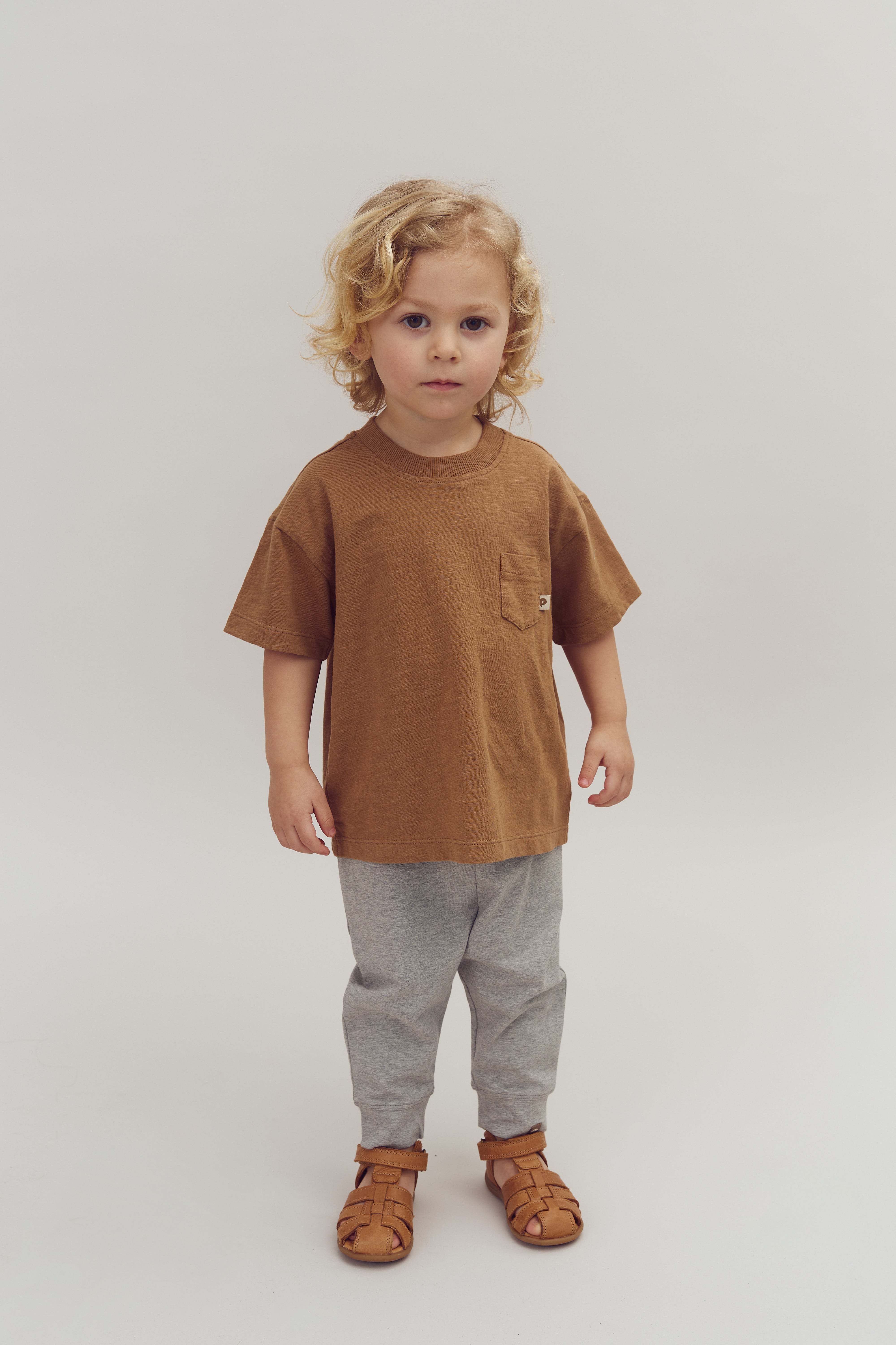 Et barn står stille iført en brun T-shirt, grå bukser og brune sandaler, midt i en enkelt hvid baggrund.