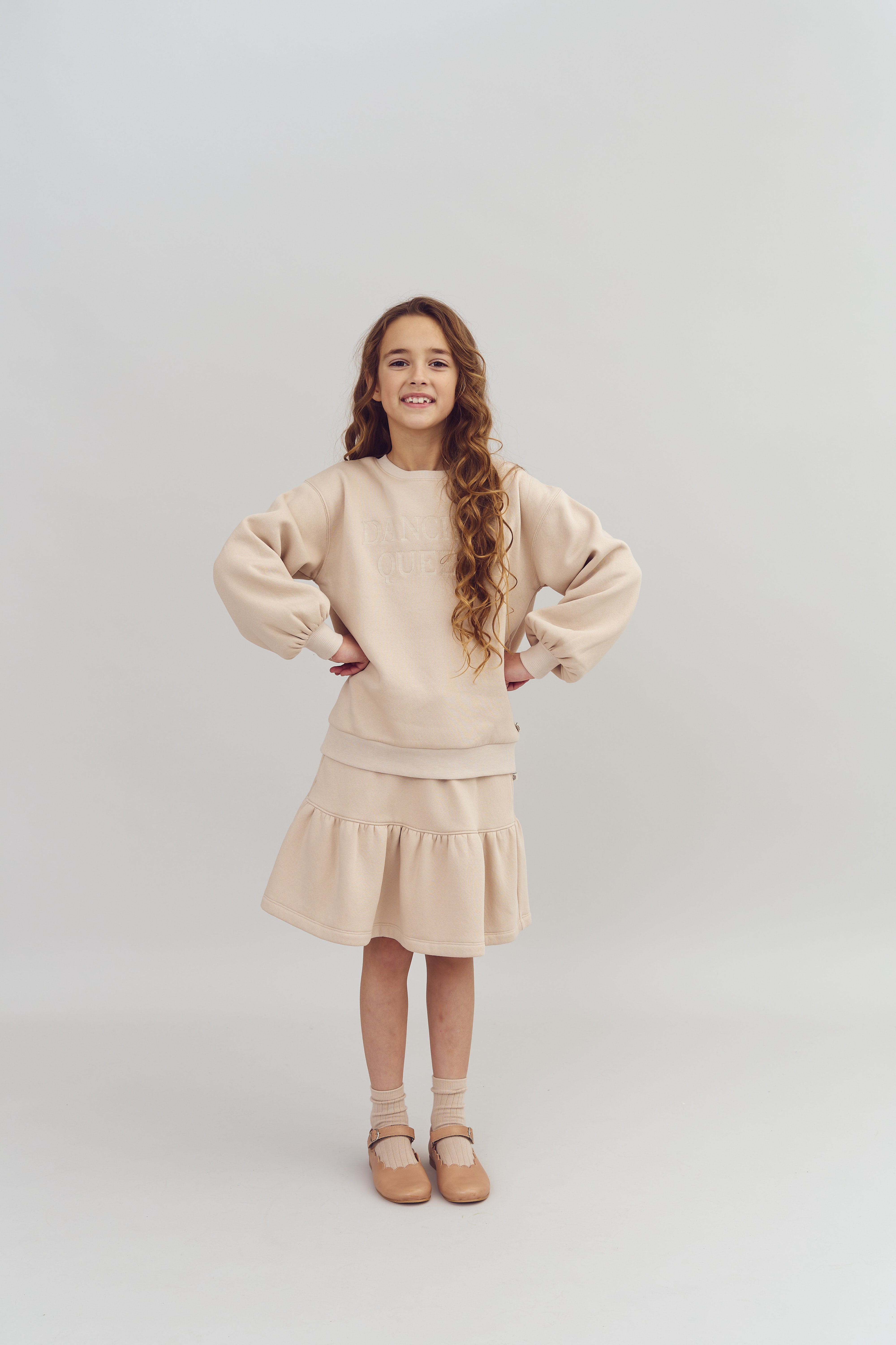 En pige står med hænderne på hofterne iført en beige trøje med teksten "DANCE QUEEN" og en nederdel, i et neutralt, enkelt studie.