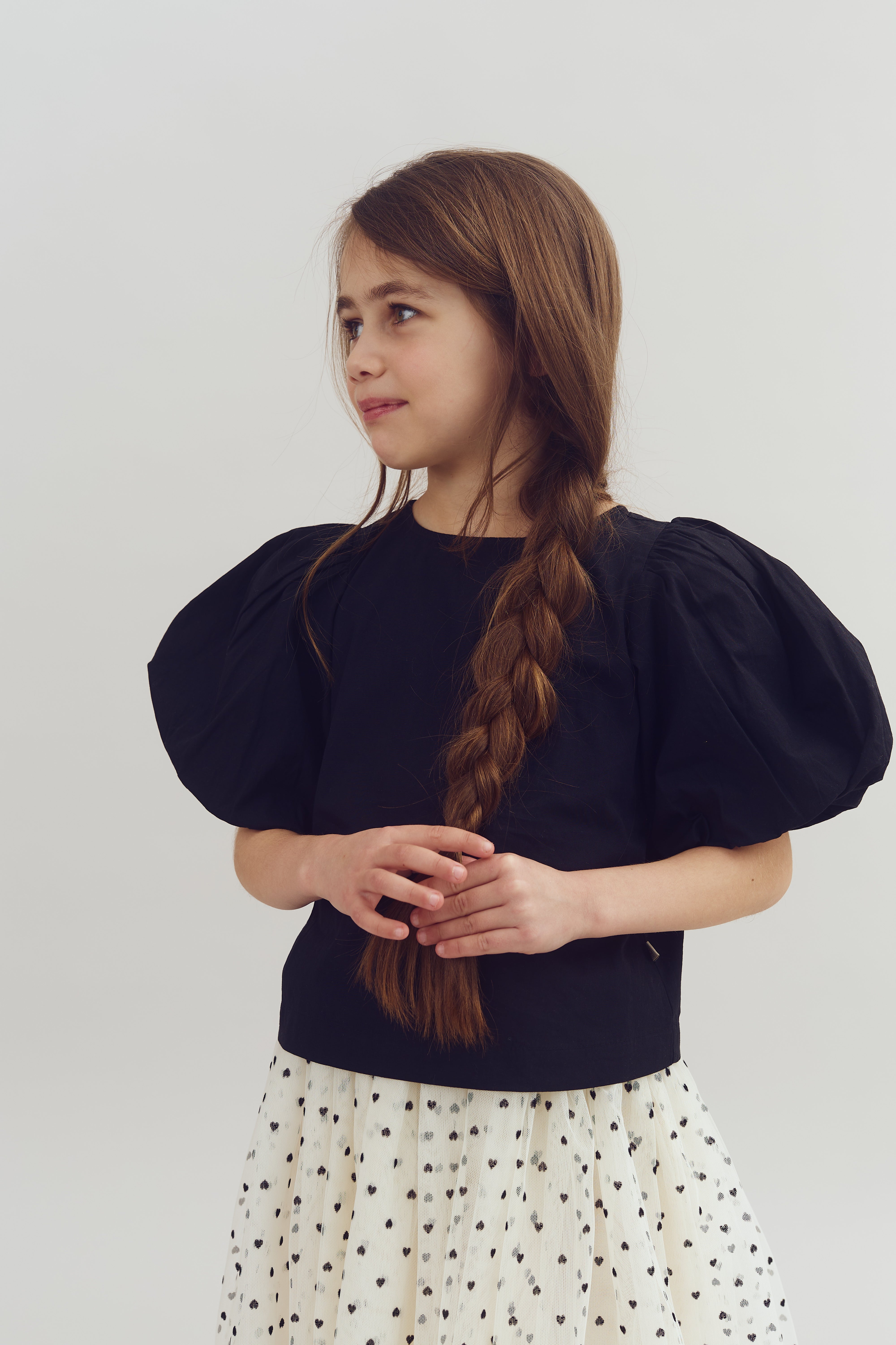A young girl stands with a slight smile, wearing a black top with puffed sleeves and a white polka-dot skirt. Her long hair is braided, set against a plain background.