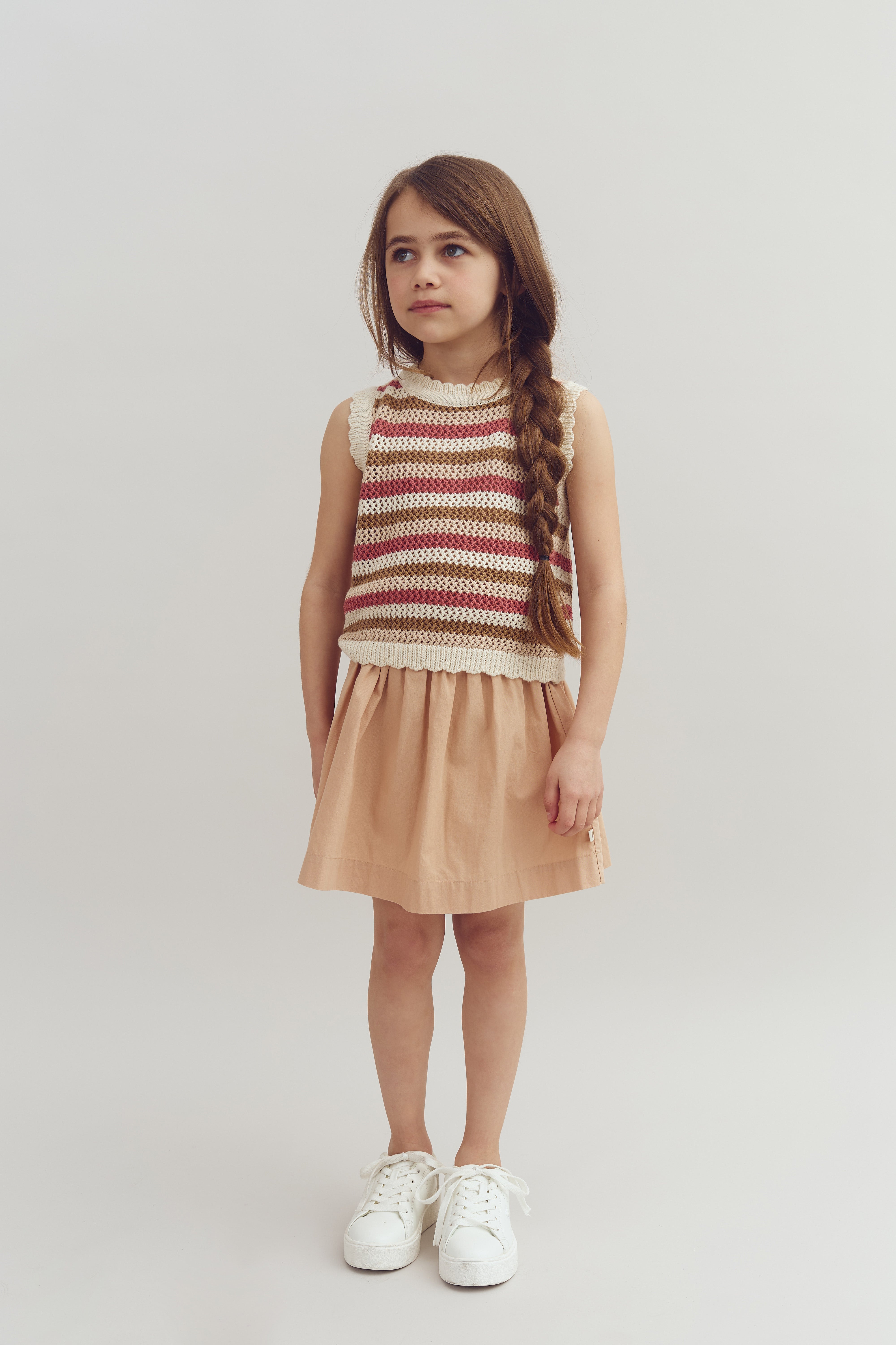 A young girl stands, wearing a striped sleeveless top and peach skirt, with a long braided hairstyle. She looks slightly to the side, set against a plain white background.
