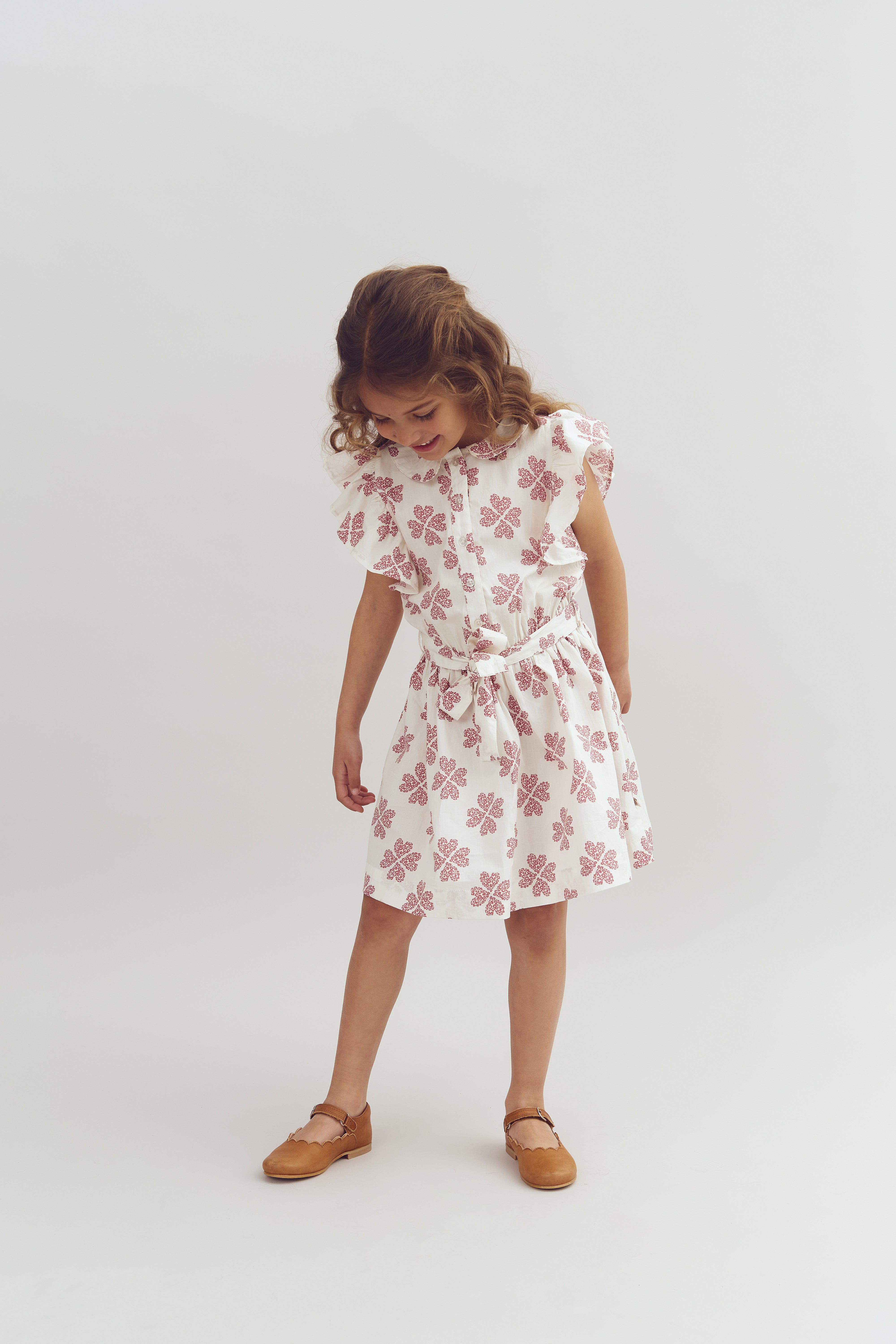 A child wearing a white dress with pink floral patterns is looking down, standing in a minimalistic, light-gray studio setting. She pairs the dress with light brown shoes.
