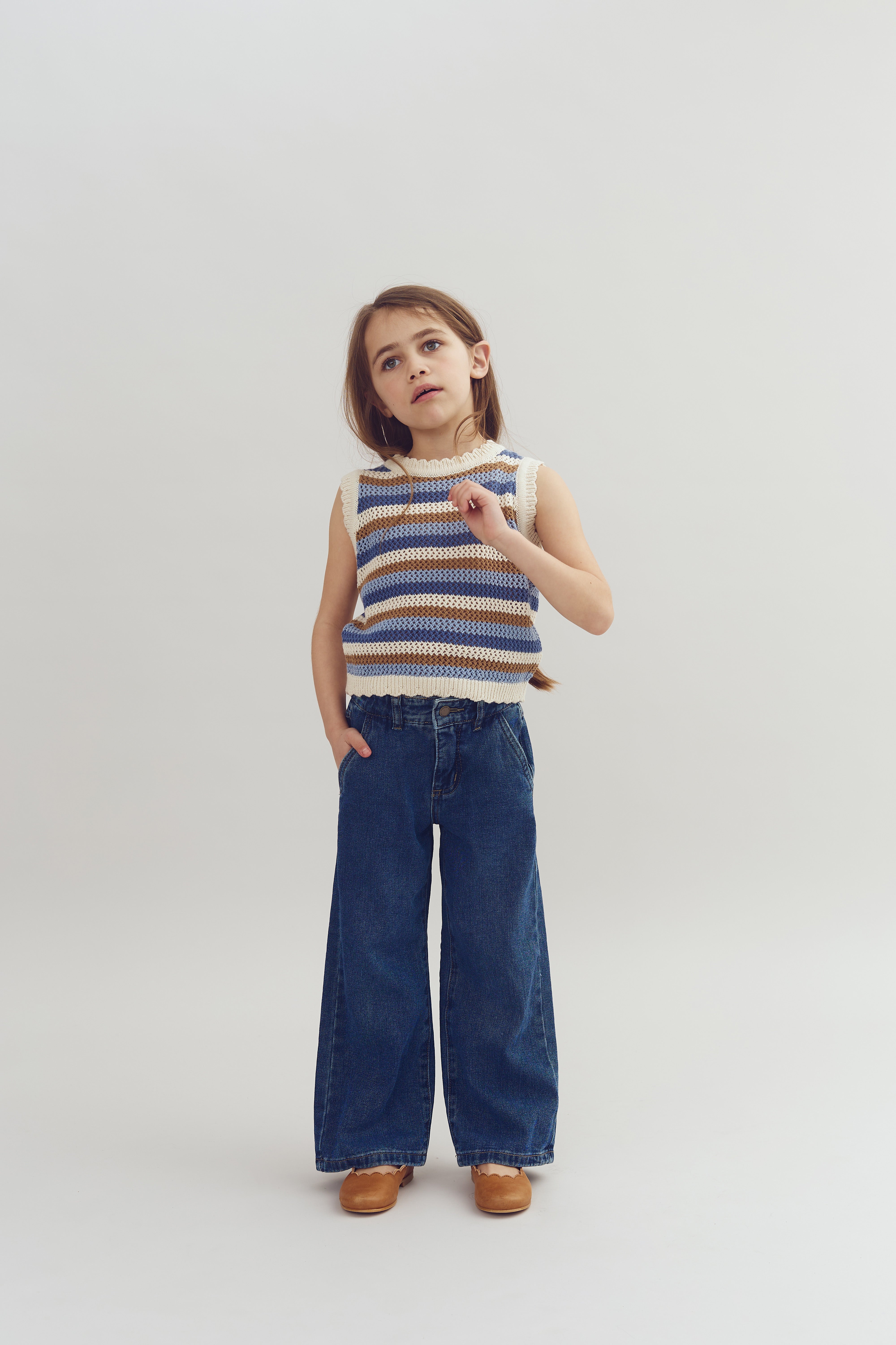 A child stands with one hand in a pocket and the other raised, wearing a striped sleeveless top and wide-leg jeans, against a plain, light background.