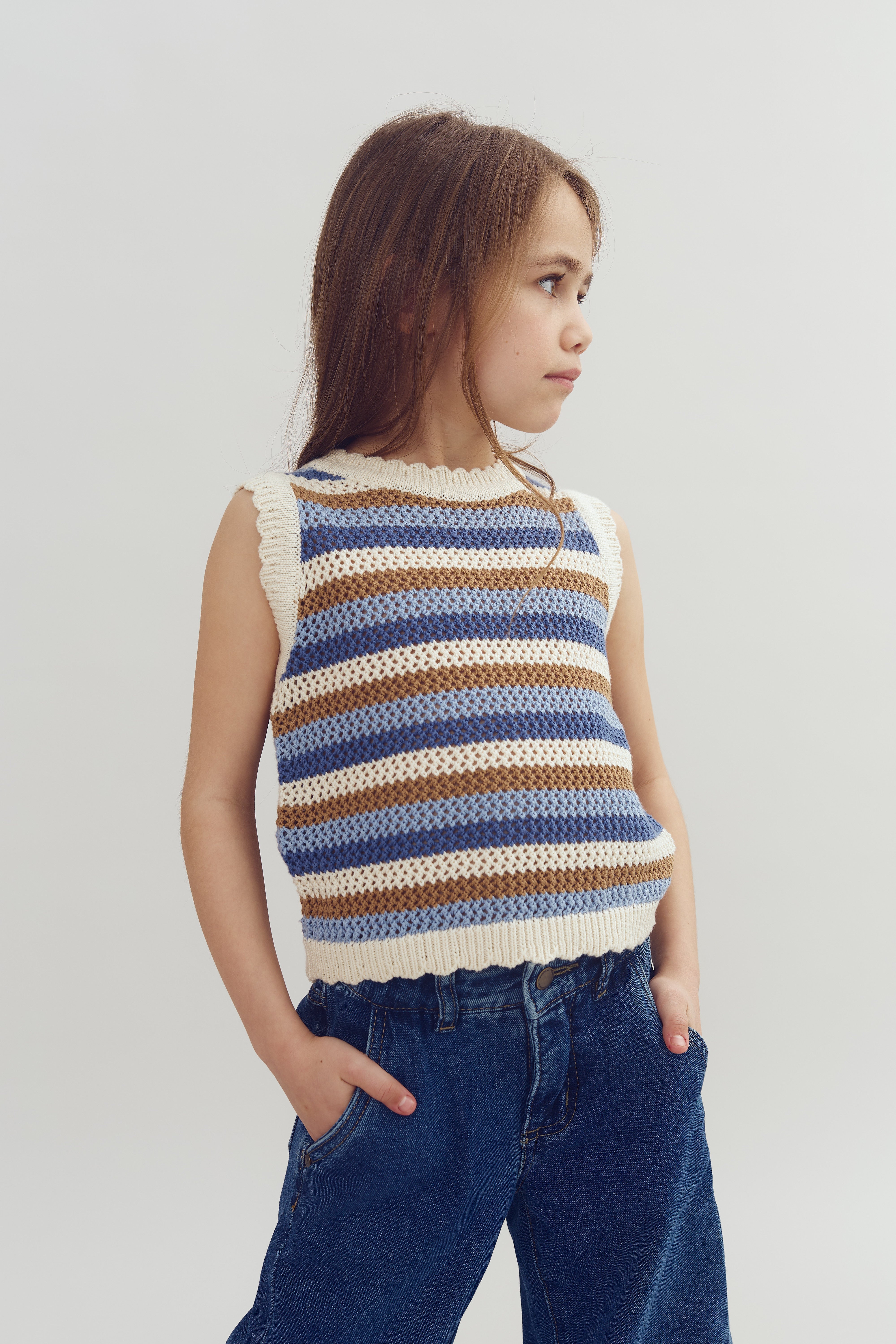 Child wearing a striped sleeveless sweater and blue jeans, standing with hands in pockets, looking to the side against a plain white background.