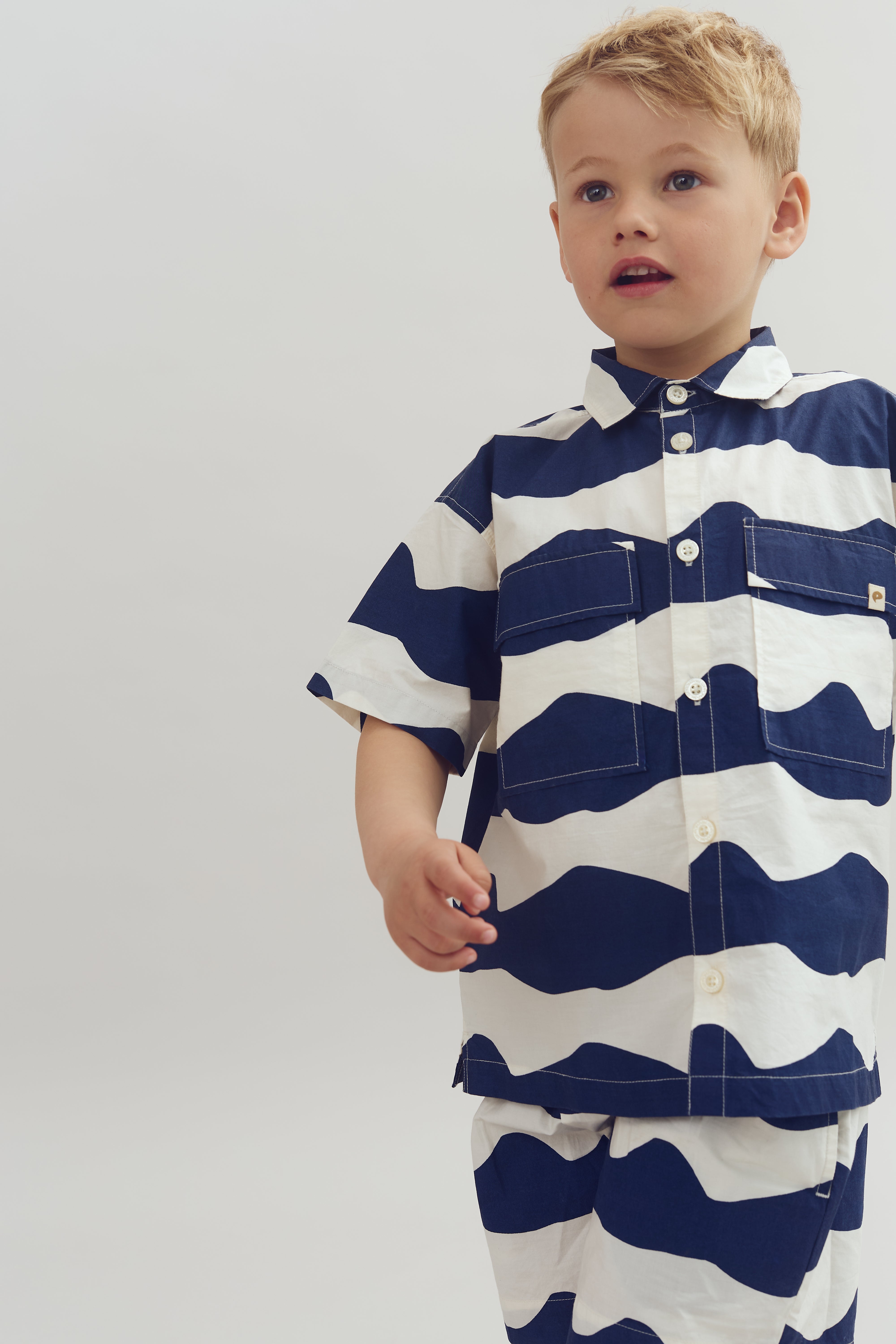 Dreng i skjorte med blå-hvide bølger står afslappet i et neutralt rum, ser opmærksom ud og holder en hånd fremme. Ingen tekst til stede.