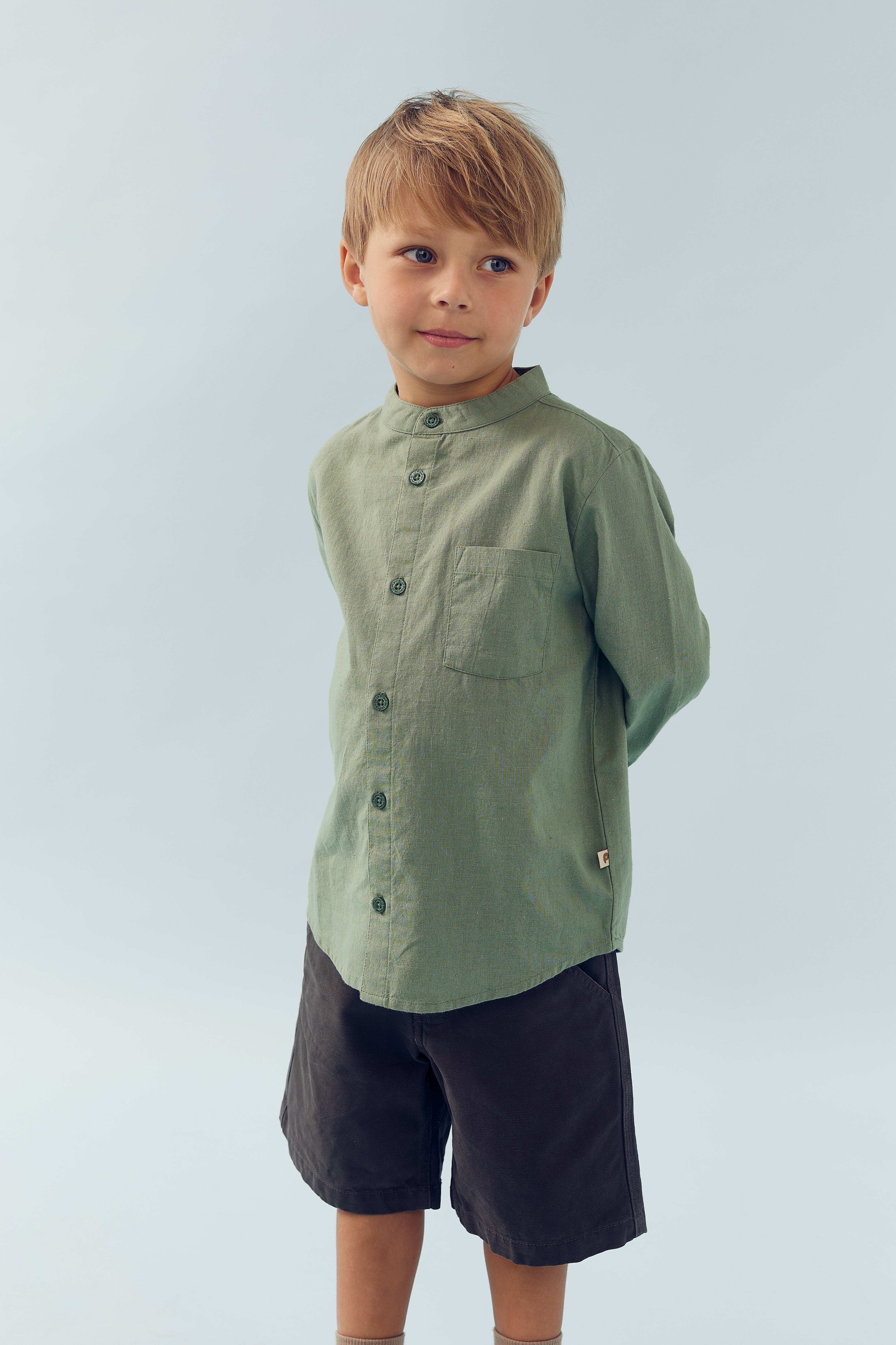 A young child stands with hands behind their back, wearing a green button-up shirt and dark shorts, against a plain light gray background.