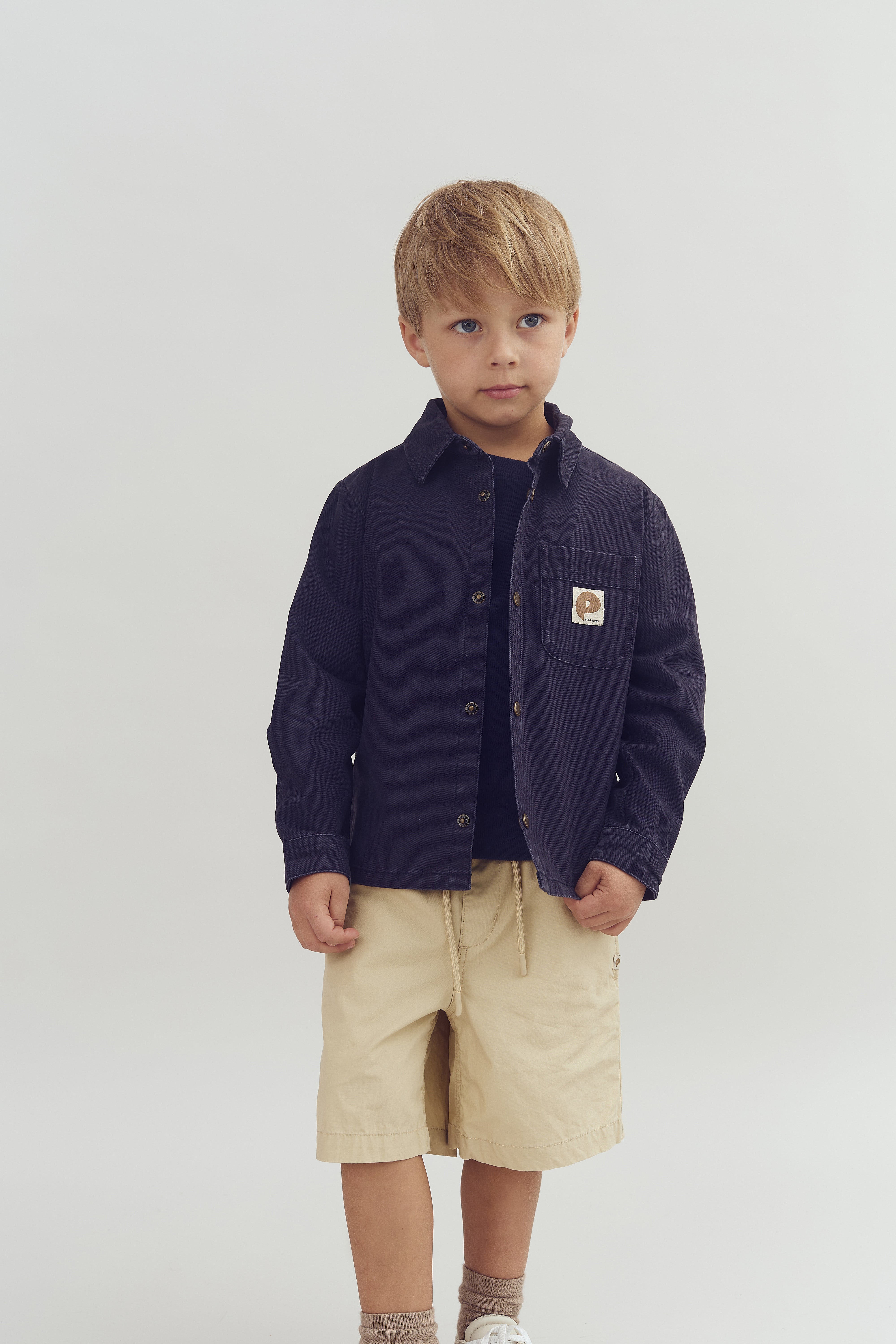 Dreng står stille iført mørkeblå skjorte med beige logo lomme og beige shorts, hvide sneakers; baggrund er hvid.