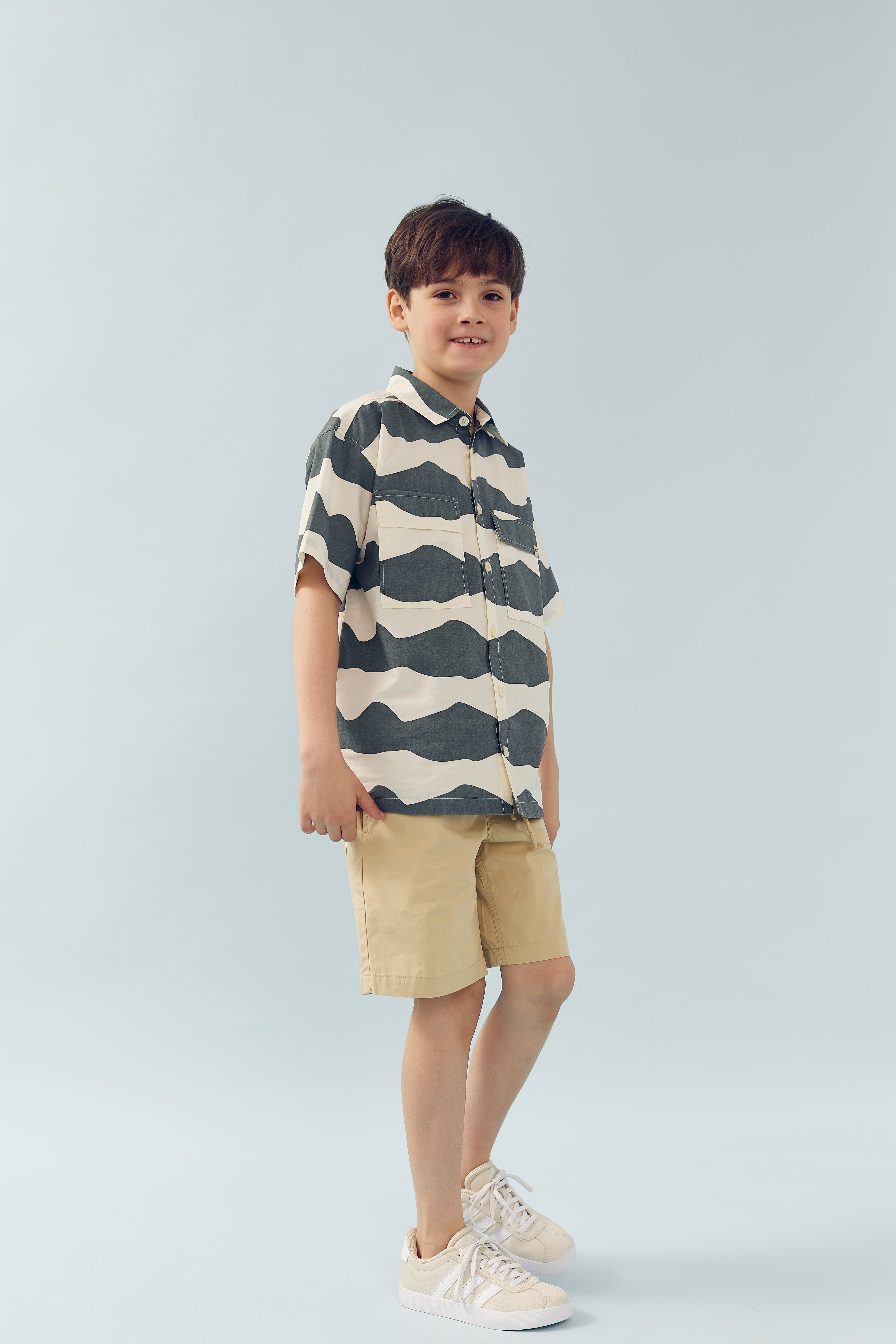 Dreng står roligt iført en mønstret skjorte, beige shorts og hvide sneakers. Baggrunden er lyseblå og minimalistisk, hvilket skaber en neutral og ren atmosfære.