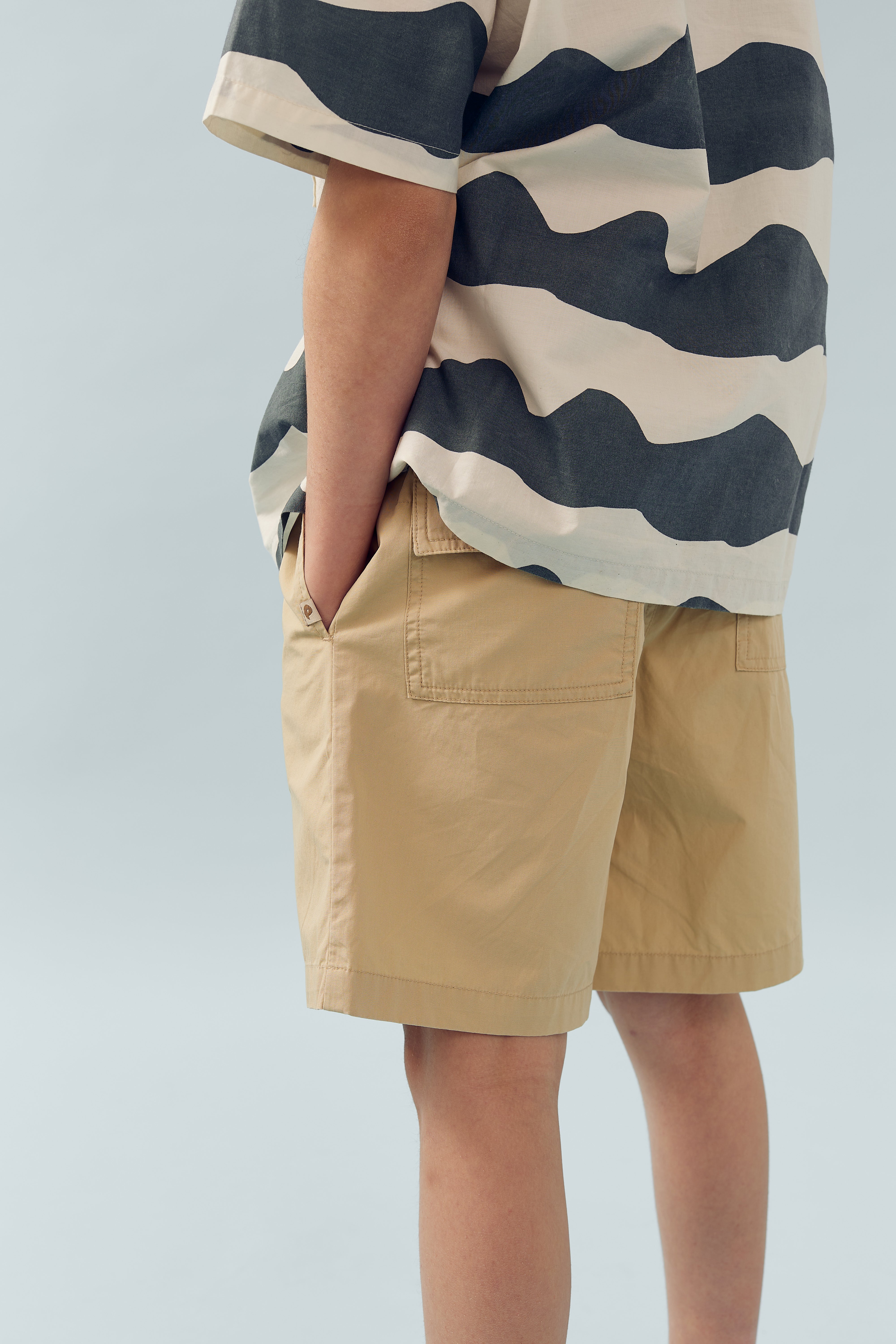 En person iført en bølge-mønstret skjorte og beige shorts står med hænderne i lommerne mod en lys baggrund.