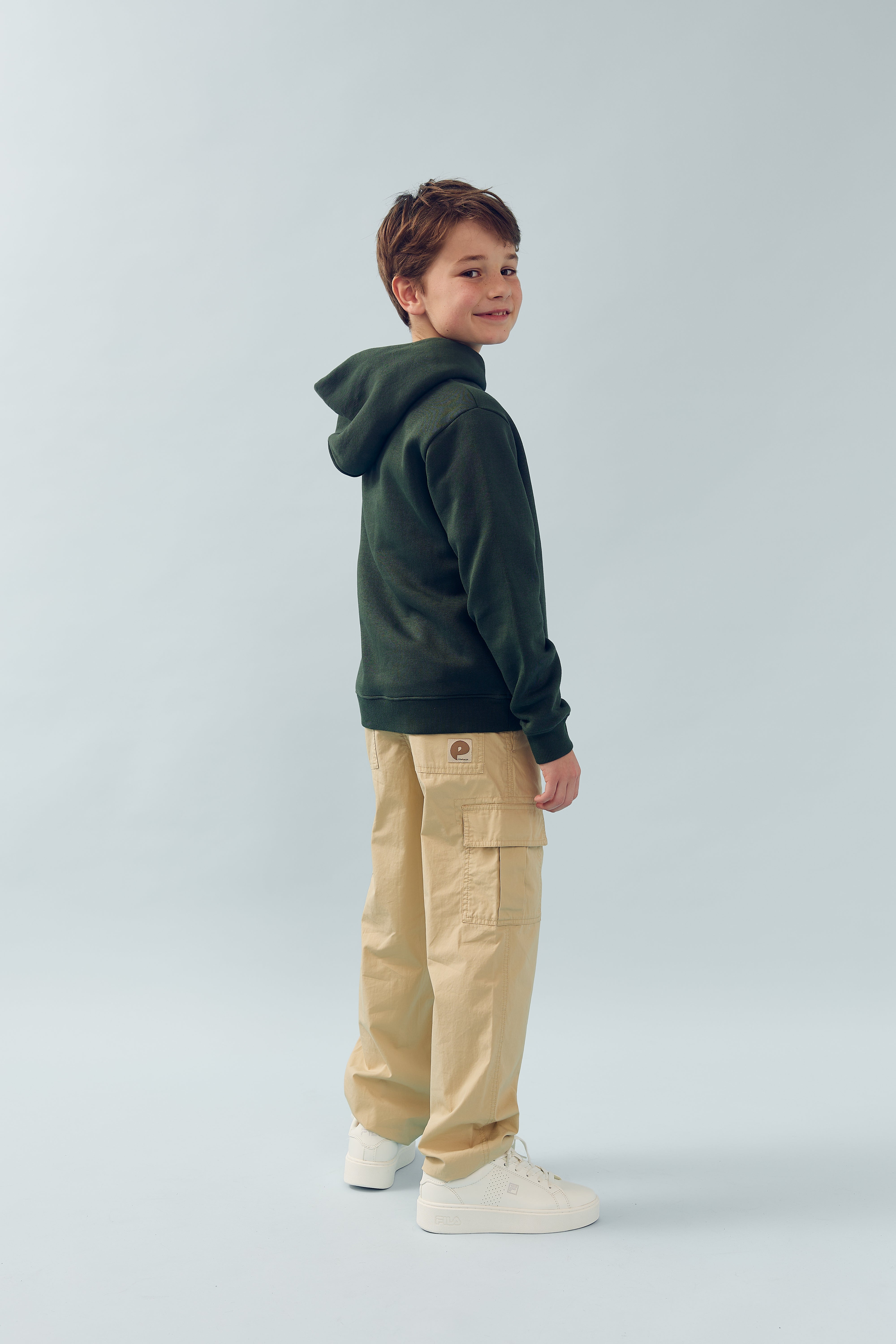 Dreng smiler, iført mørkegrøn hættetrøje og beige cargo-bukser, står i profil. Baggrund er en lys, ensfarvet væg.