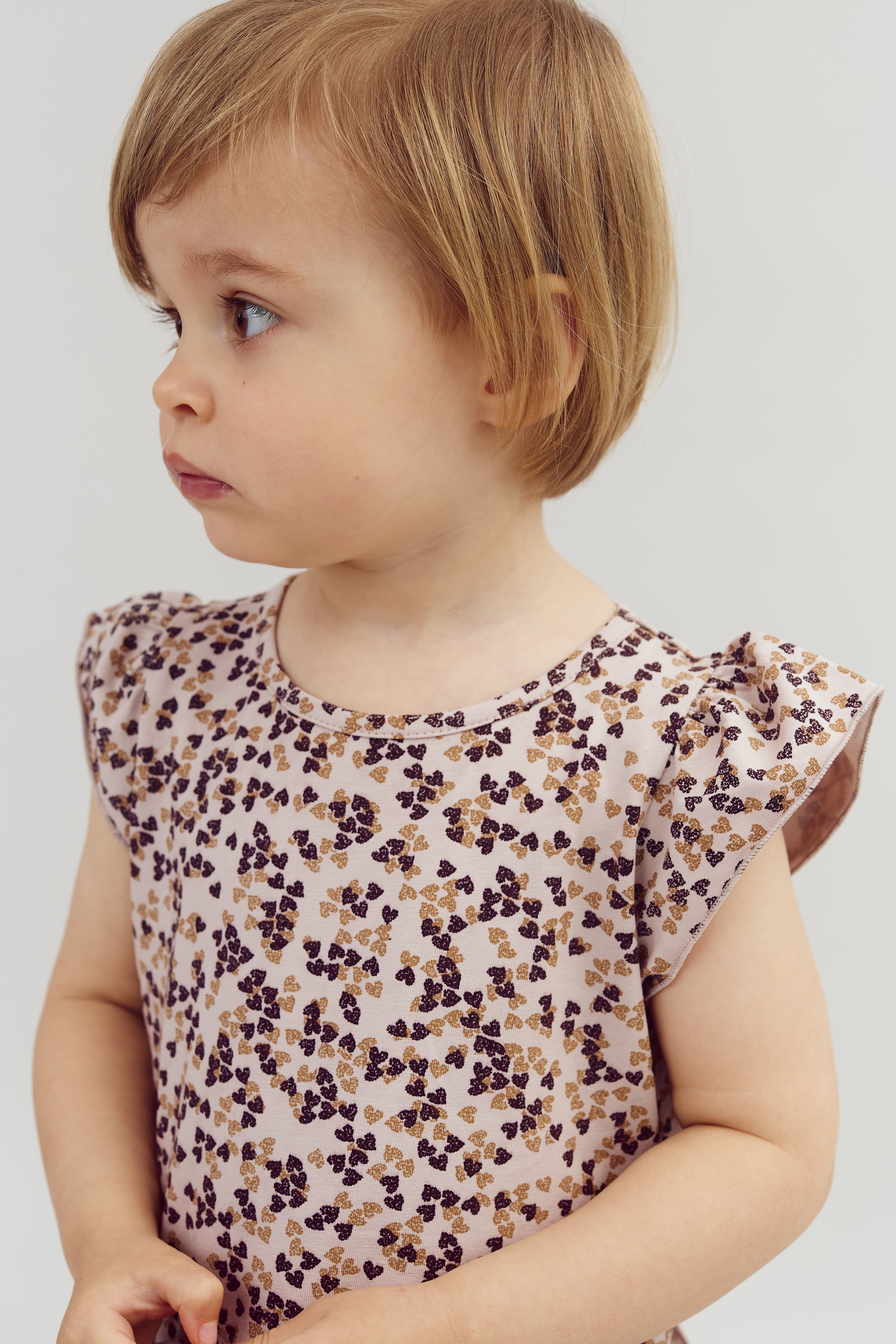 A young child, looking sideways, is wearing a patterned shirt with small brown and purple shapes, in a neutral, softly lit setting.