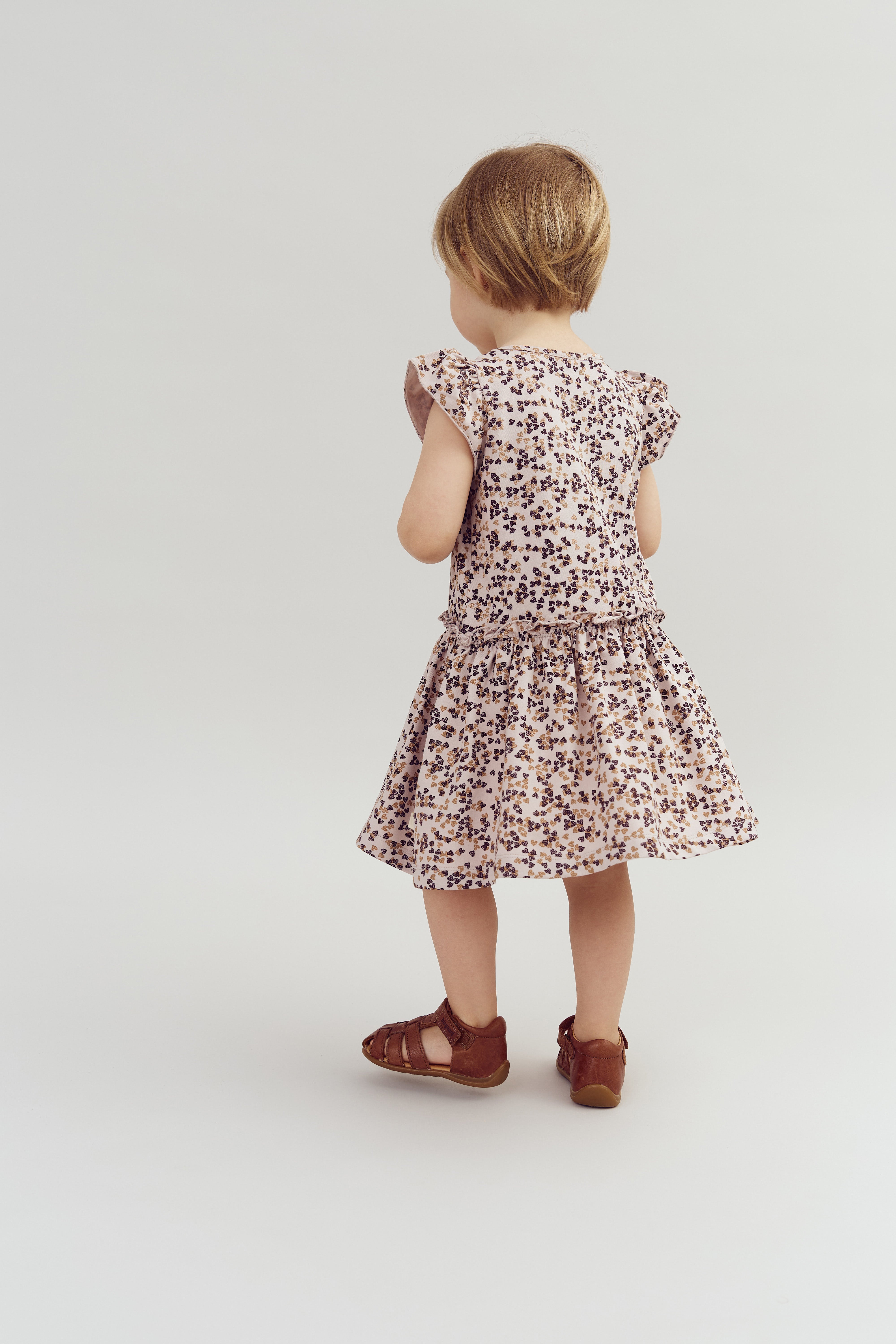 A child stands, facing away, wearing a floral-patterned dress and brown sandals, on a light, neutral background.