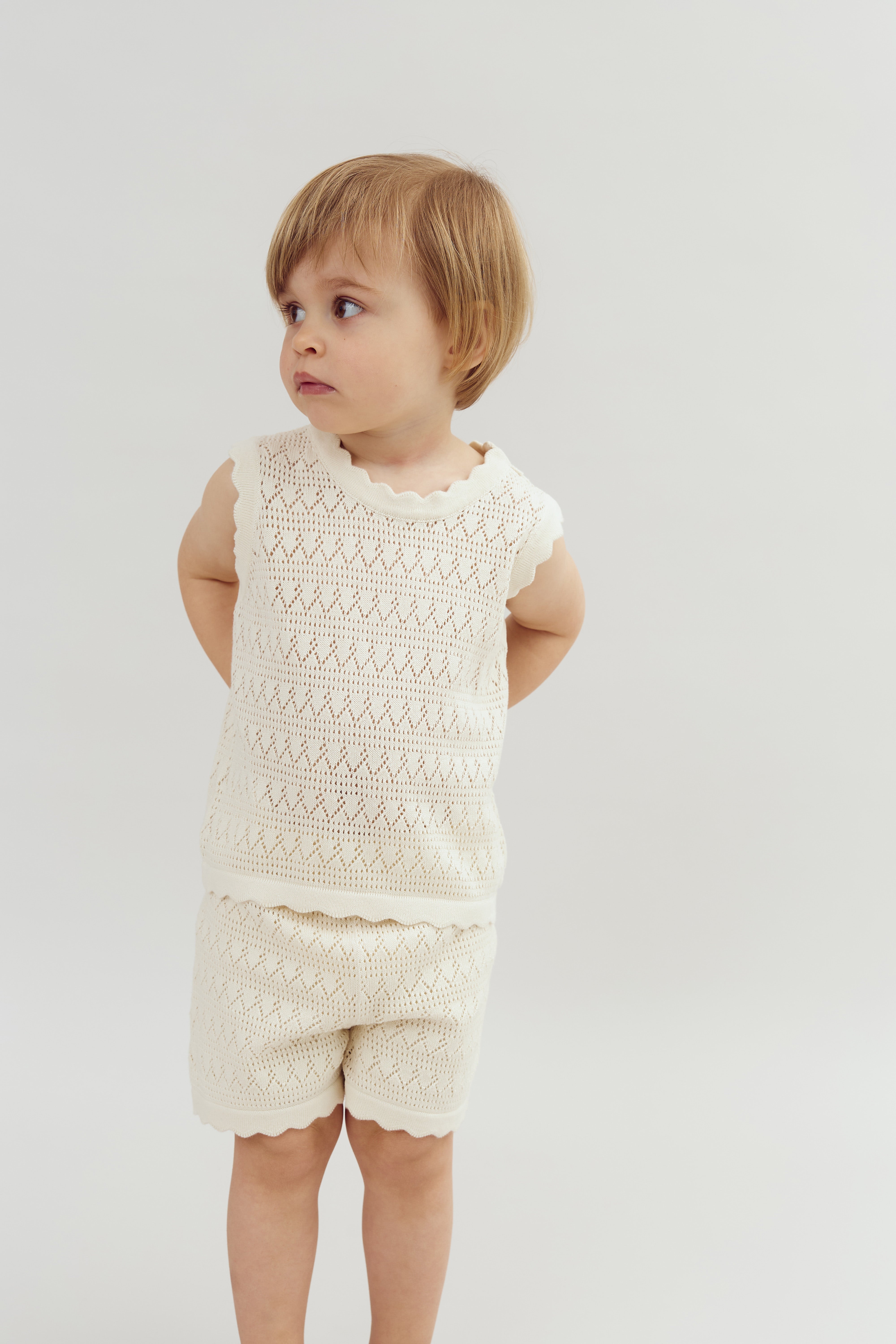 A young child stands with hands behind their back, wearing a knitted cream outfit, looking to the side. They are set against a plain, light-colored background.