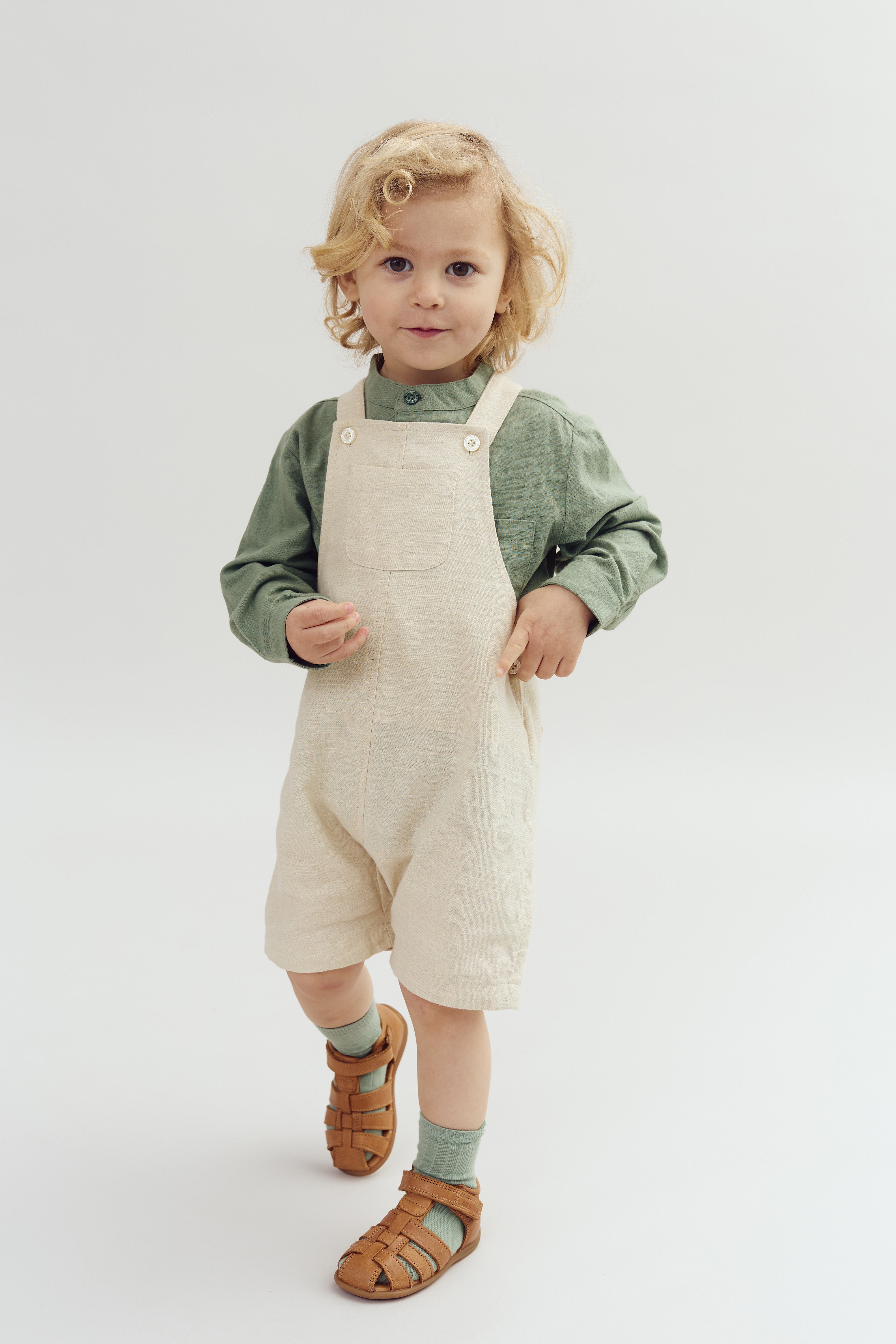 Et lille barn med lyst, krøllet hår står iført en grøn bluse og lyse overalls, iført brune sandaler, på en ensfarvet, lys baggrund.