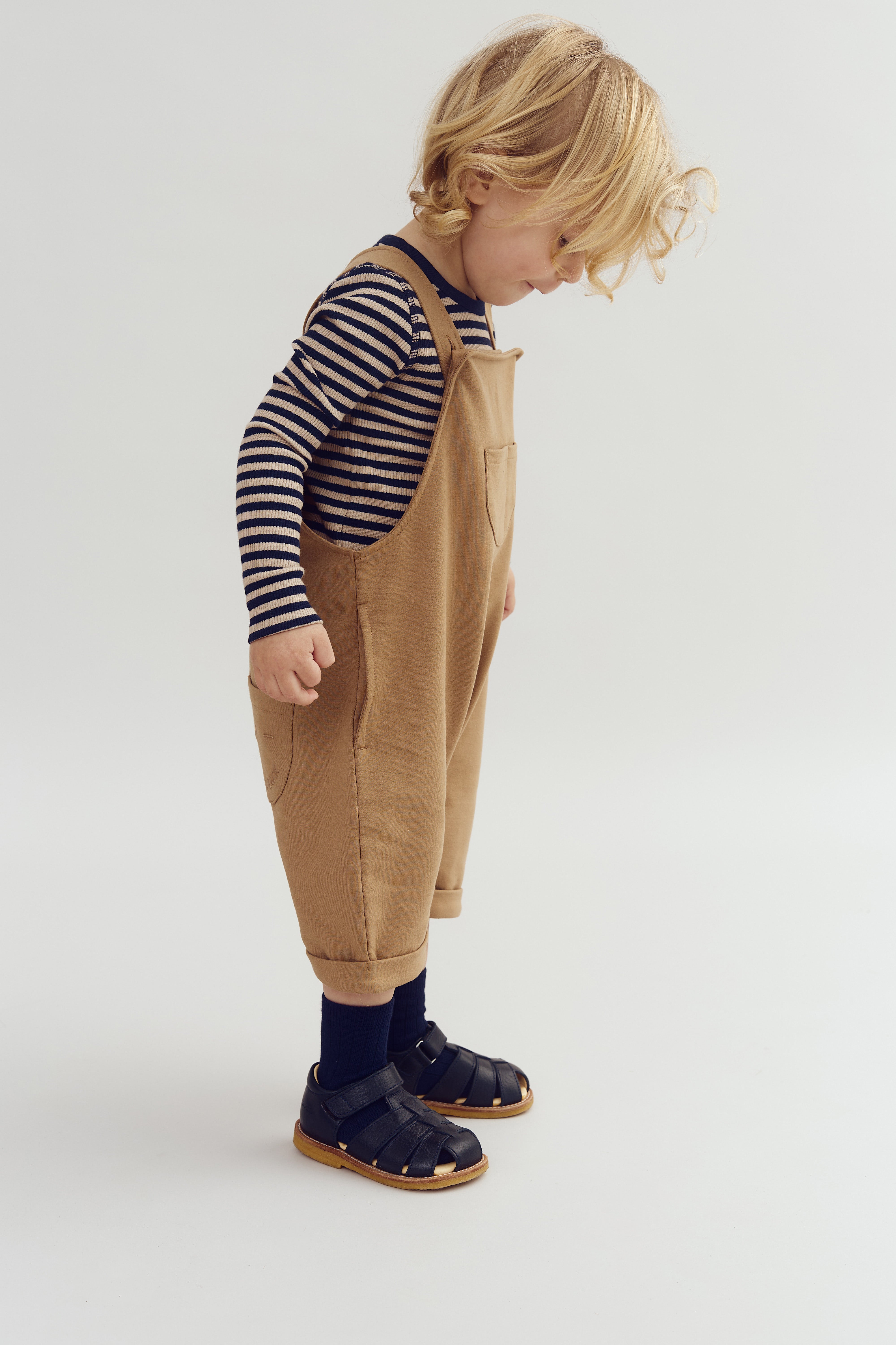 Child stands side-on, wearing tan overalls and striped long-sleeve shirt, looking down. They are in black sandals with socks, on a plain white background.