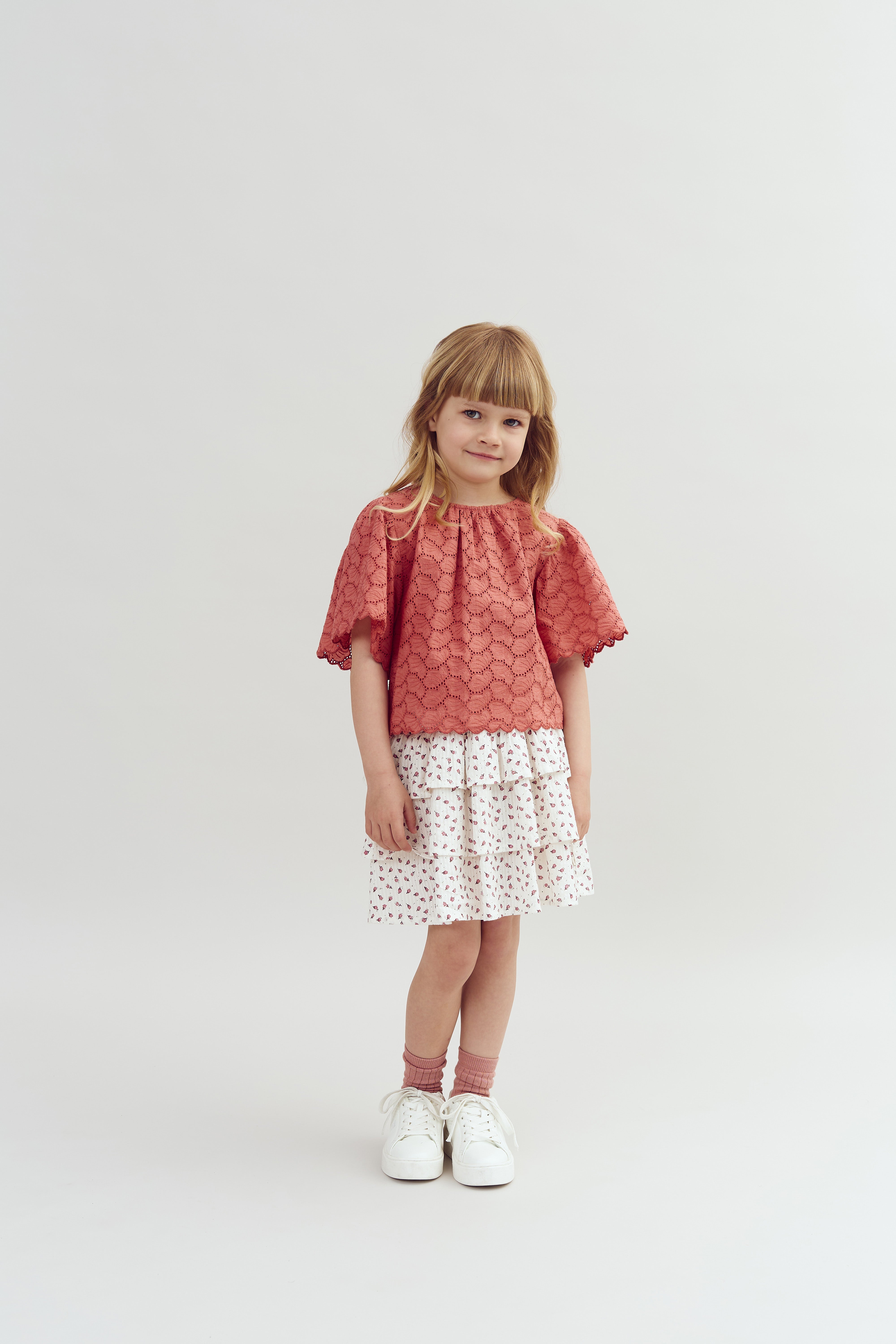 A young girl stands smiling, wearing a red lace top, tiered floral skirt, and white sneakers, with a plain, light-colored background.