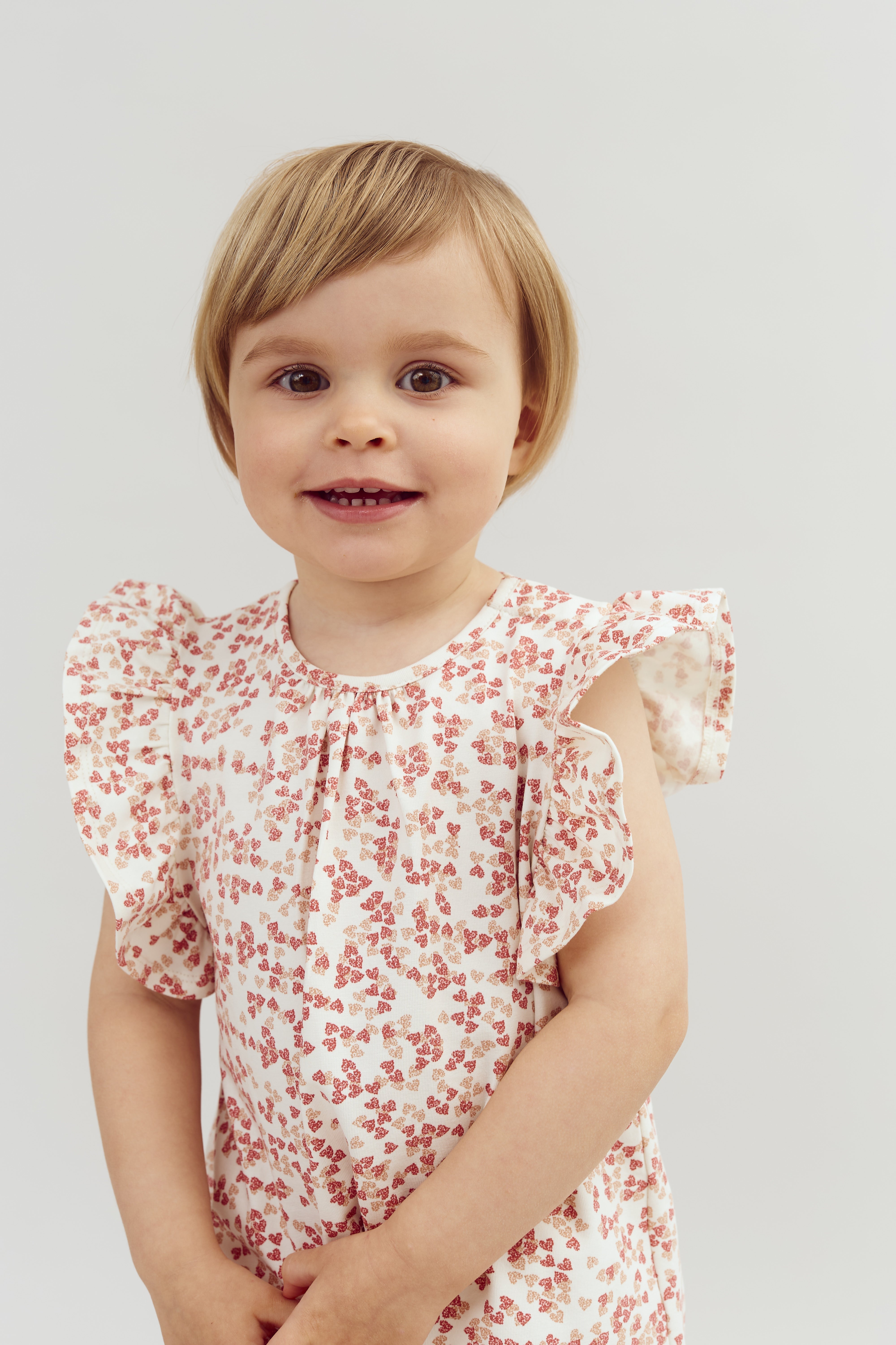Barn smiler iført en kjole med røde blomsterprint. Baggrunden er lys og enkel, hvilket fremhæver barnets udtryk og tøj.