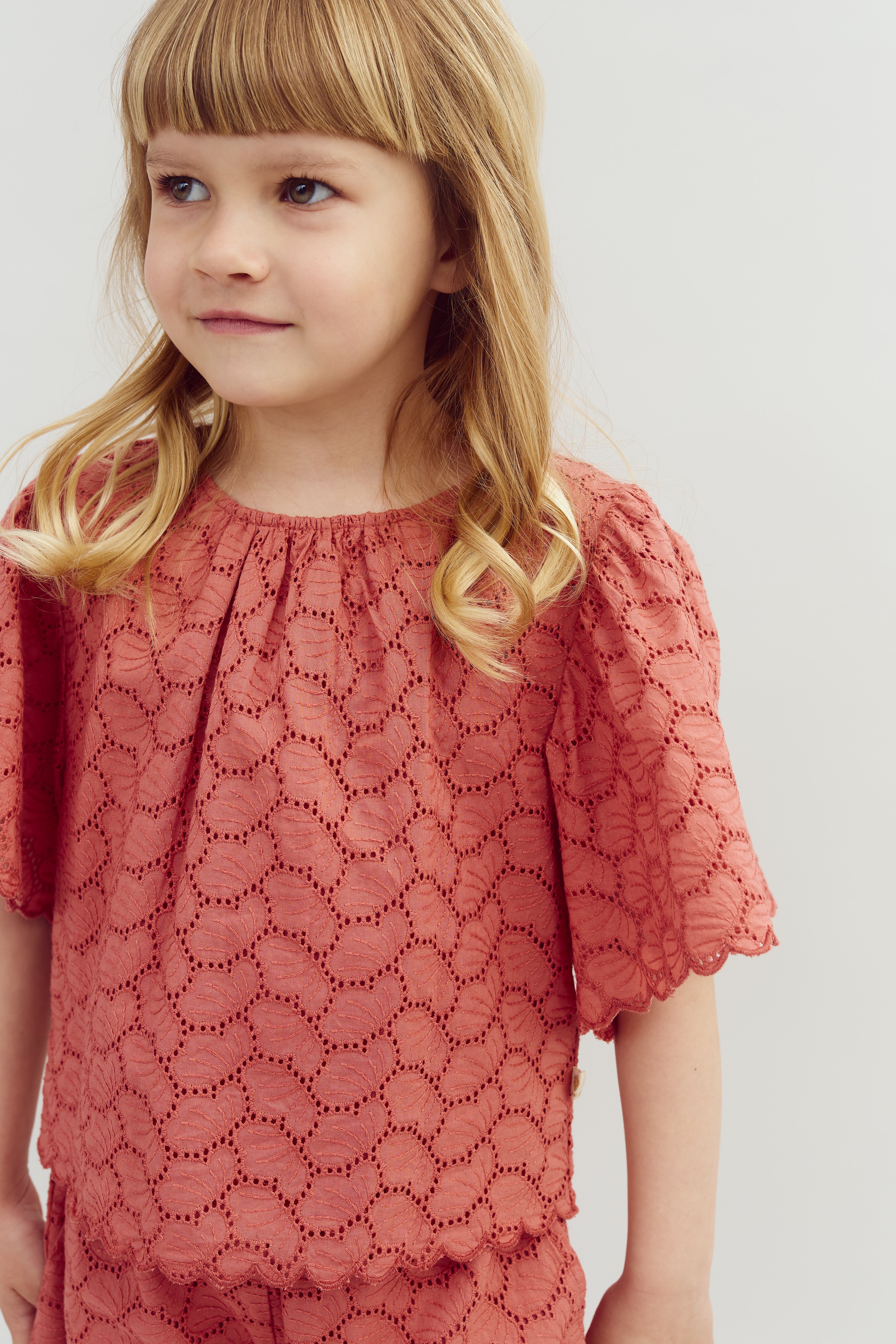 A young child with light brown hair stands, wearing a textured, pink lace top. The background is a plain, light-colored wall.