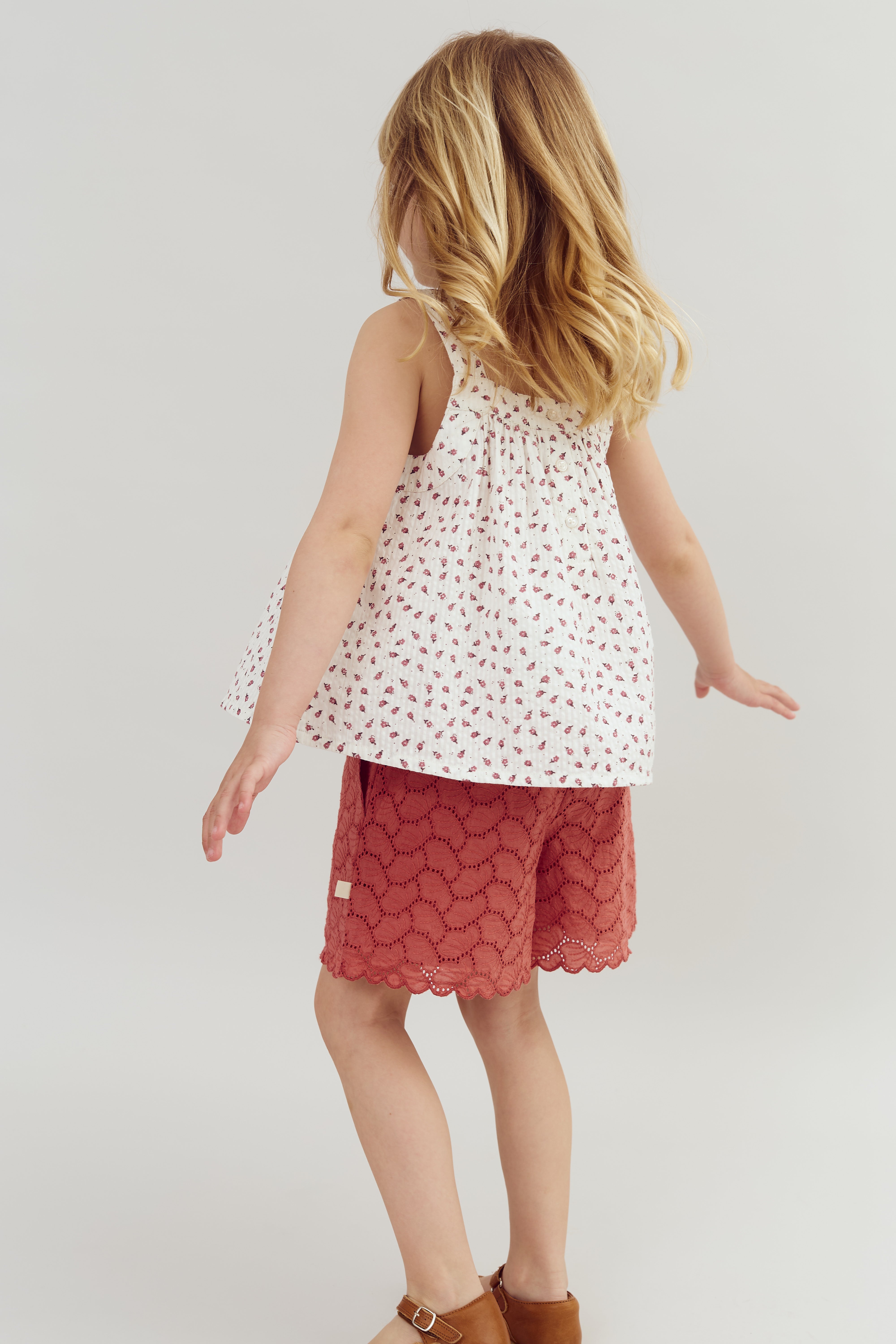 A young child with blonde hair is turning away, wearing a sleeveless white top with small red patterns and red scalloped shorts, standing against a plain background.
