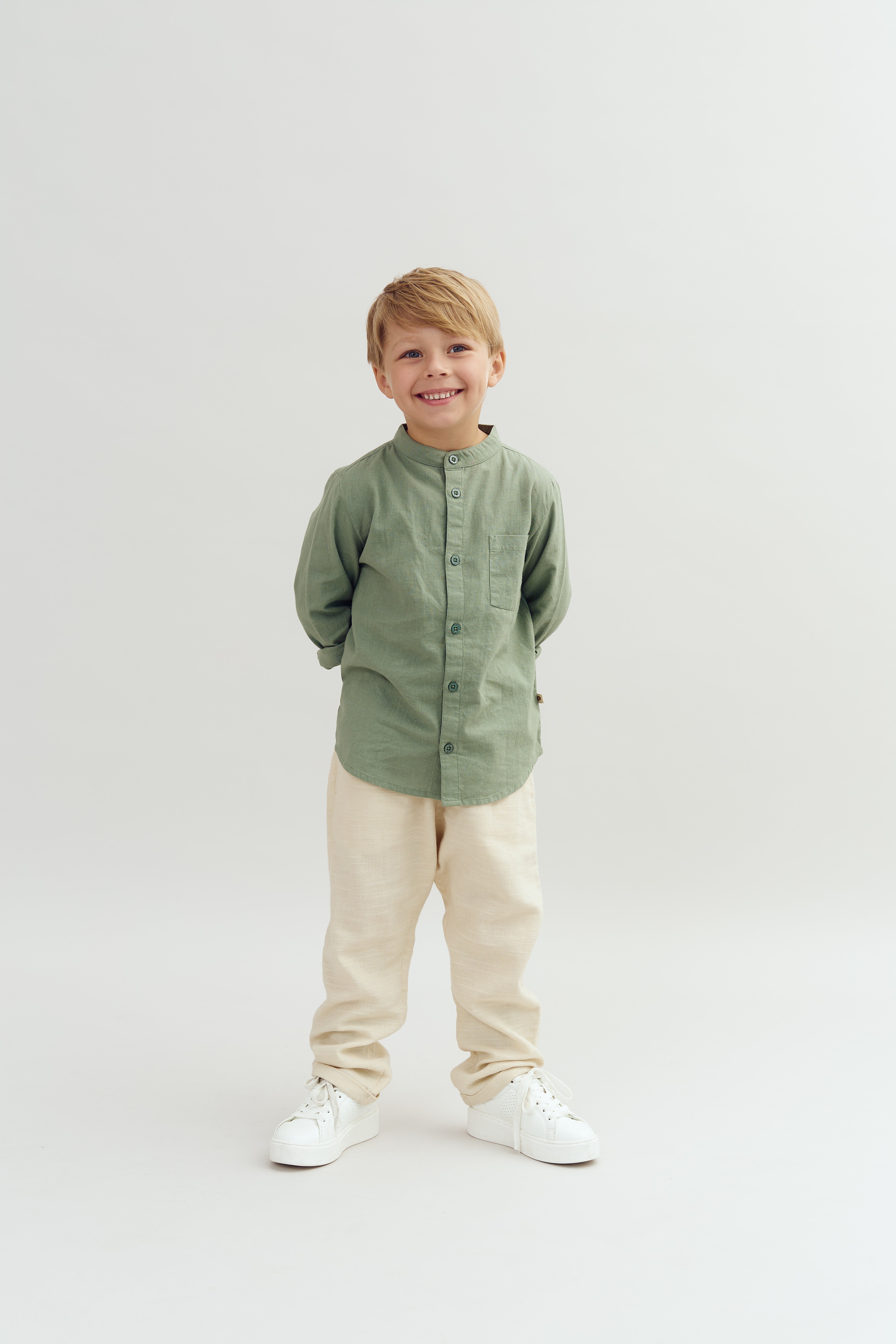 Dreng står smilende, iført en lys grøn skjorte og beige bukser, mod en neutral, lys baggrund. Han har hænderne bag ryggen og hvide sko på.