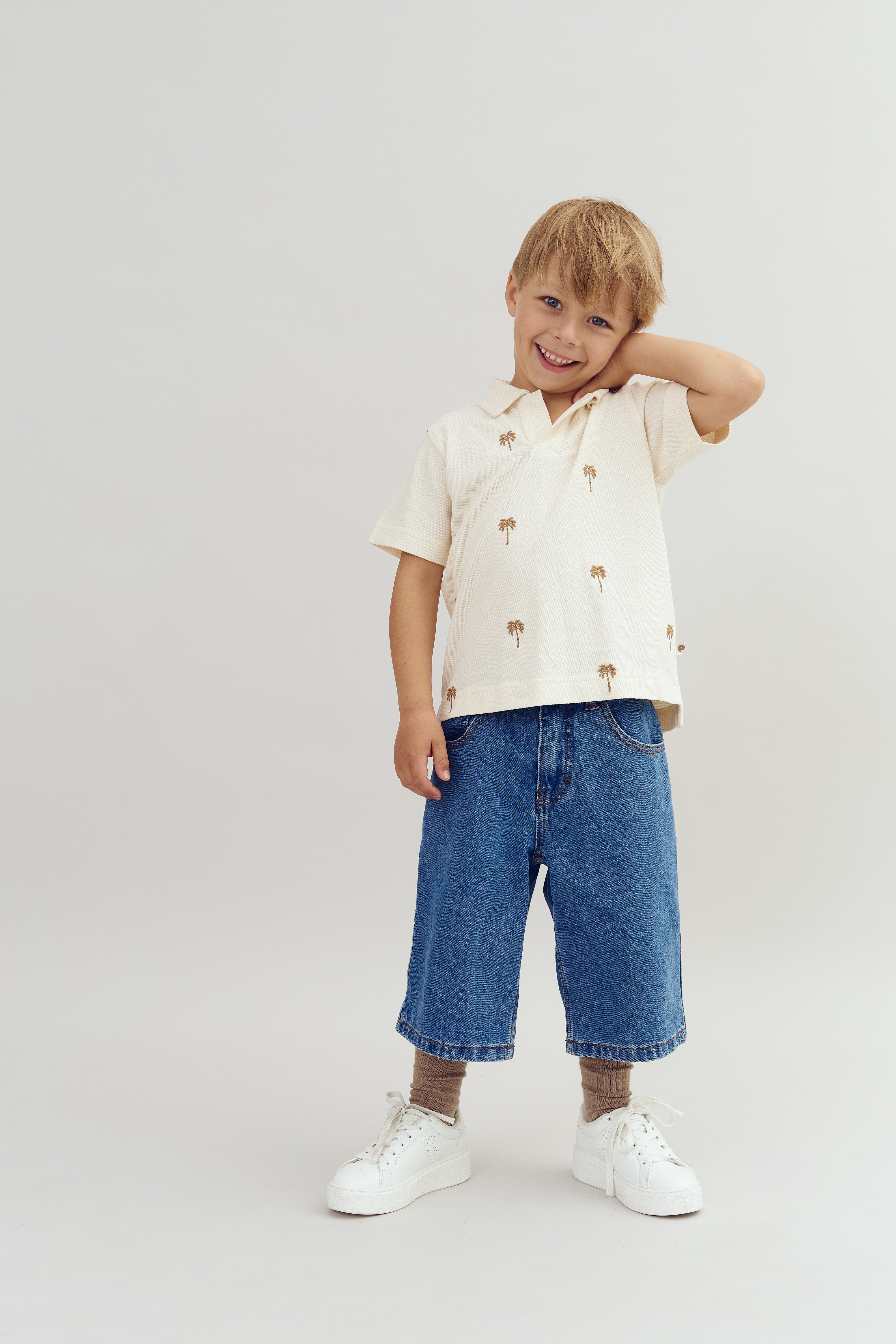 A child stands on a plain background, smiling with one hand on the neck, wearing a light shirt with palm tree designs, denim shorts, and white sneakers.