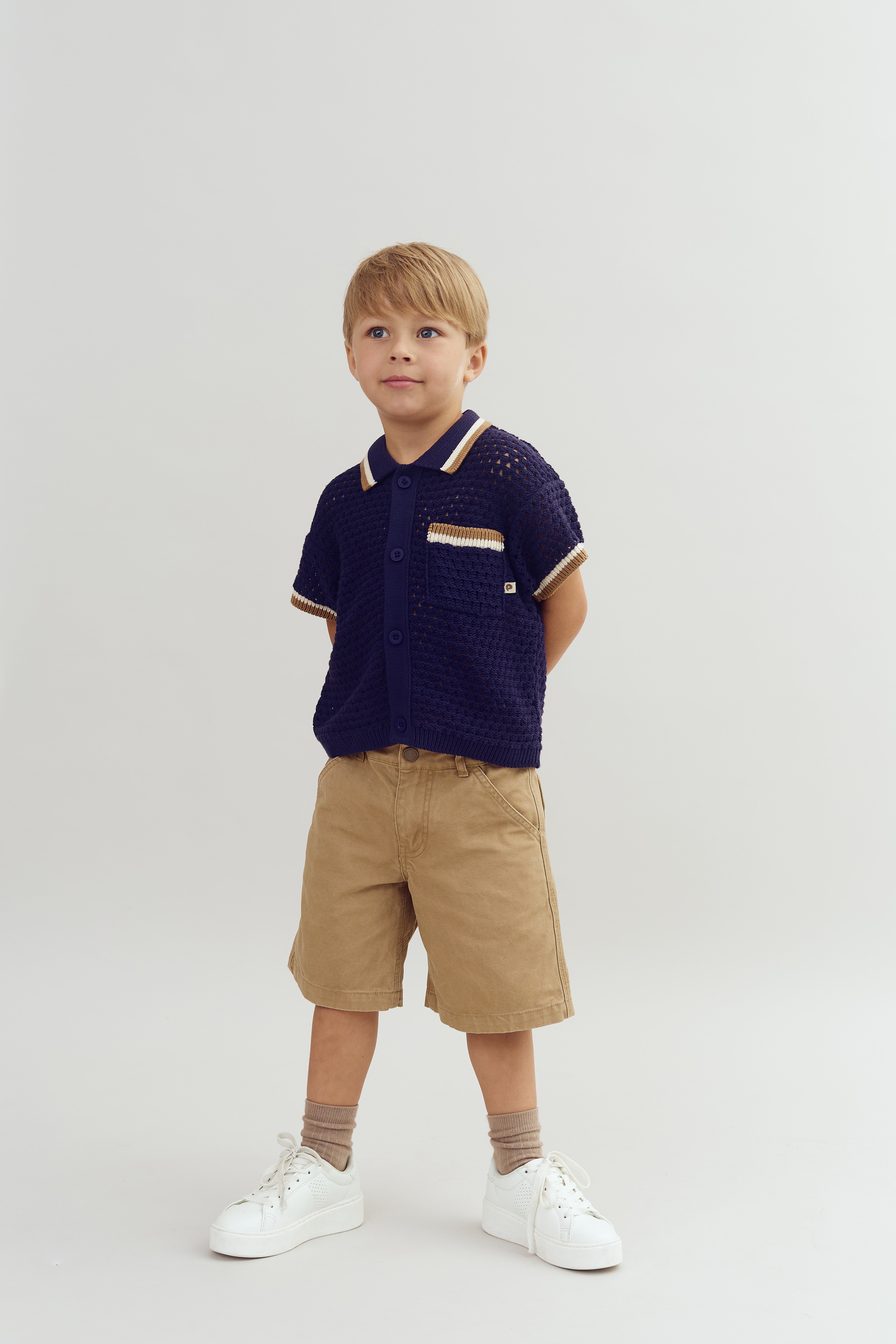 A young child stands confidently, wearing a navy blue short-sleeved shirt and beige shorts, against a plain white background. The outfit is complemented by white sneakers and socks.
