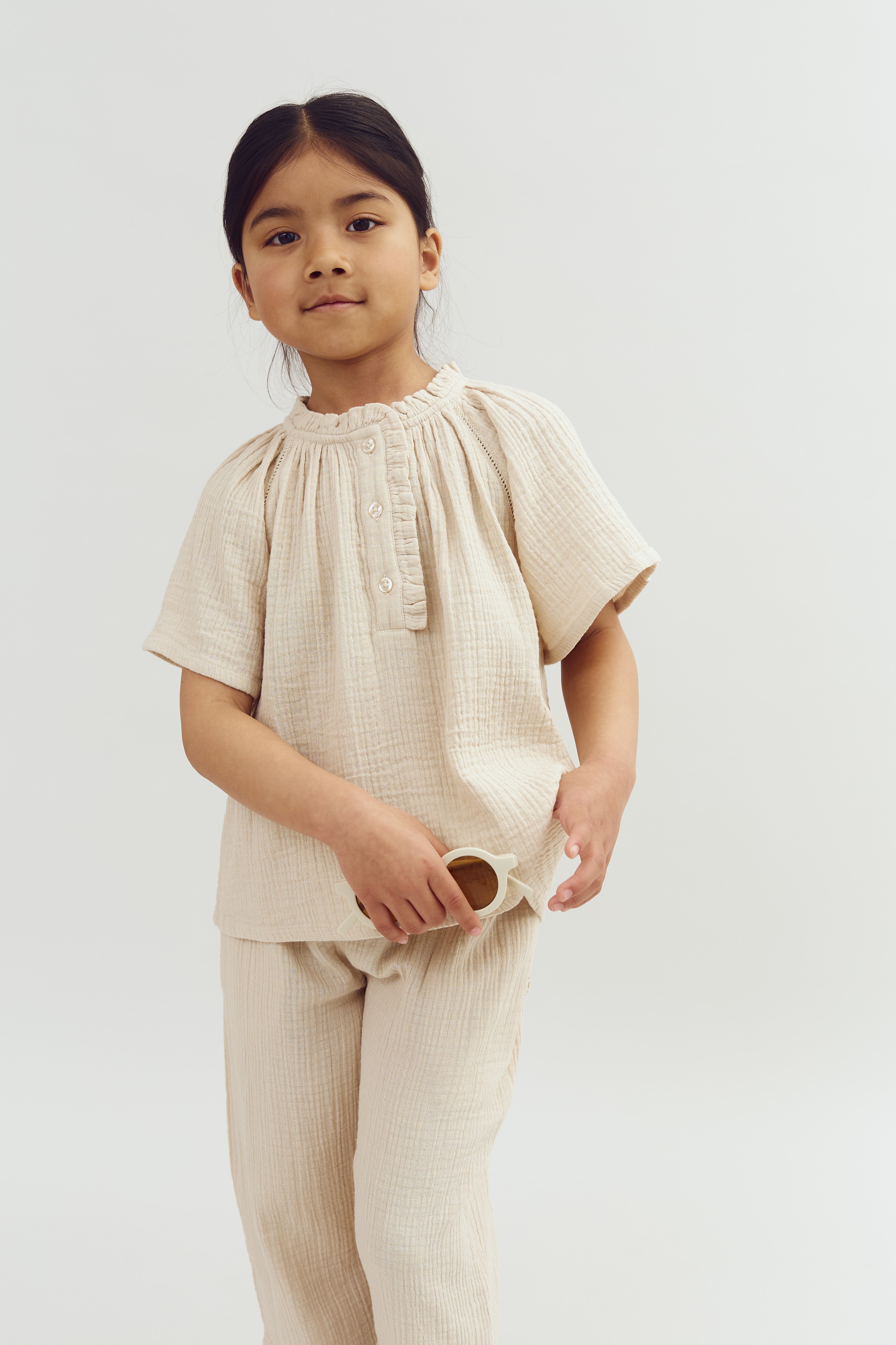 A child stands, holding sunglasses, wearing a light-textured, buttoned outfit. The background is plain white, creating a minimalist setting.