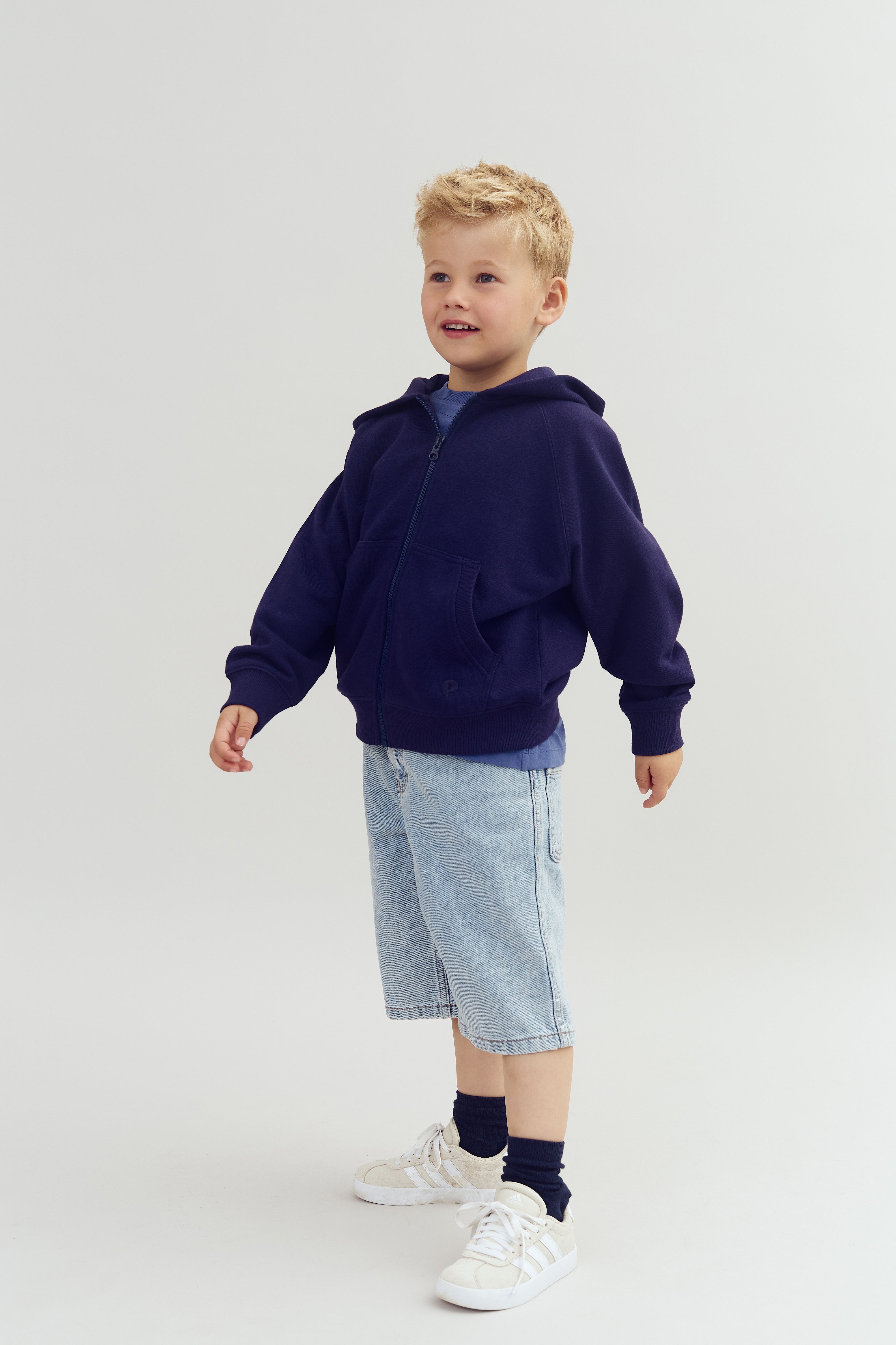 Dreng i mørkeblå hættetrøje og lyse denimshorts står og smiler. Baggrunden er enkel og lys. Han bærer hvide sneakers og mørke sokker.