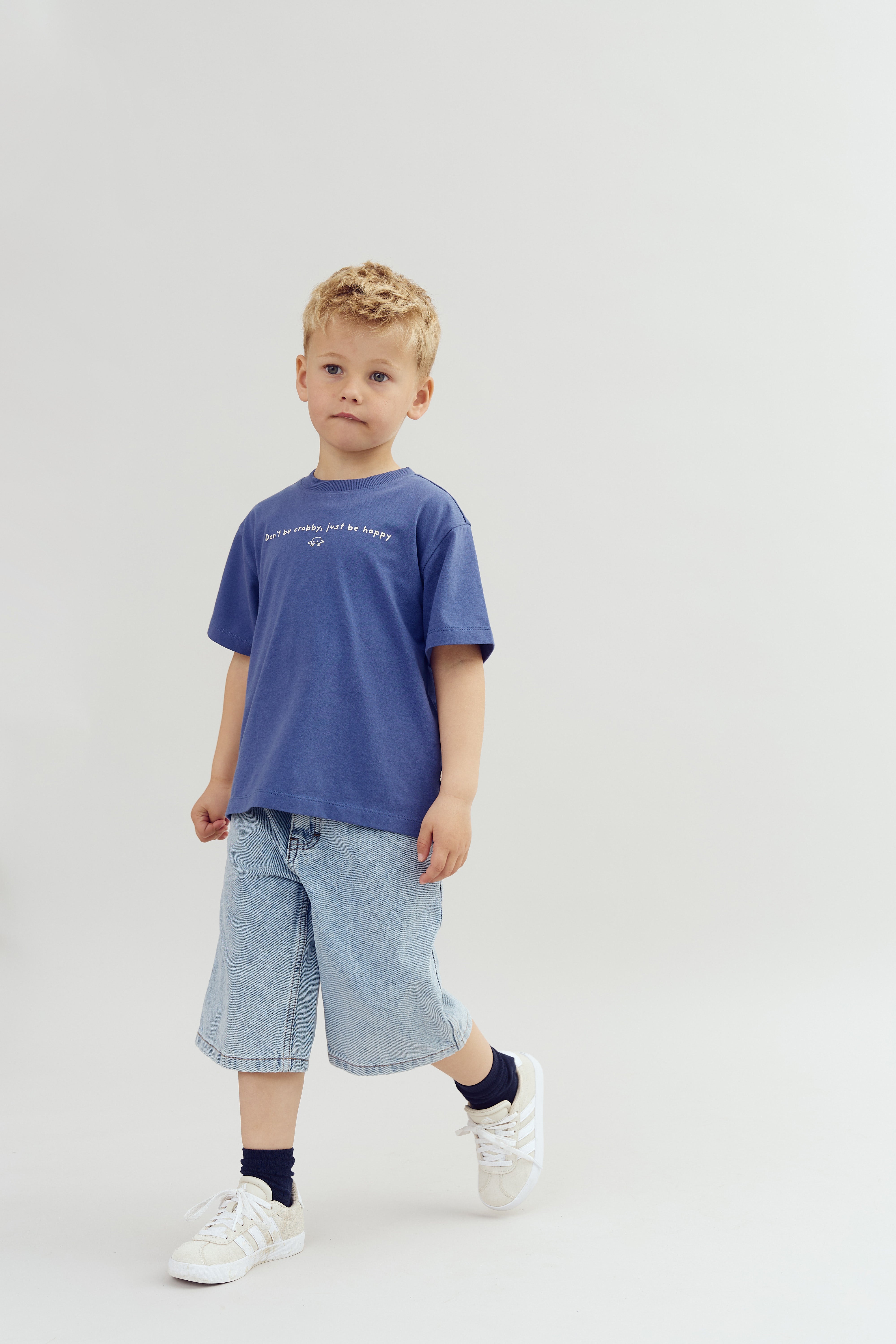 En dreng står iført en blå t-shirt og denimshorts i en lys baggrund. Der står “Don't be crabby, just be happy” på t-shirten.