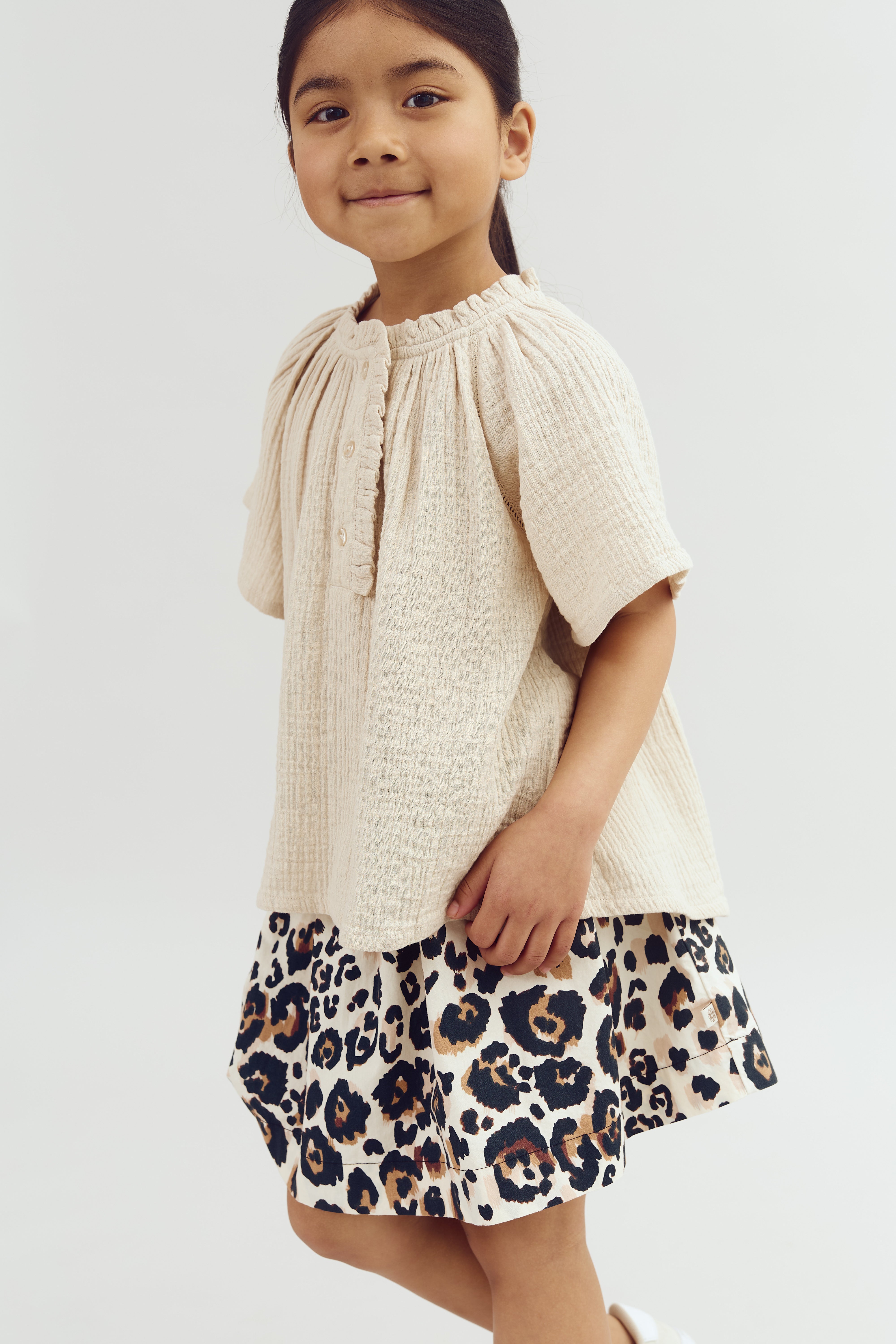 A child wears a cream-colored blouse and a leopard-print skirt, standing confidently in a neutral studio setting.