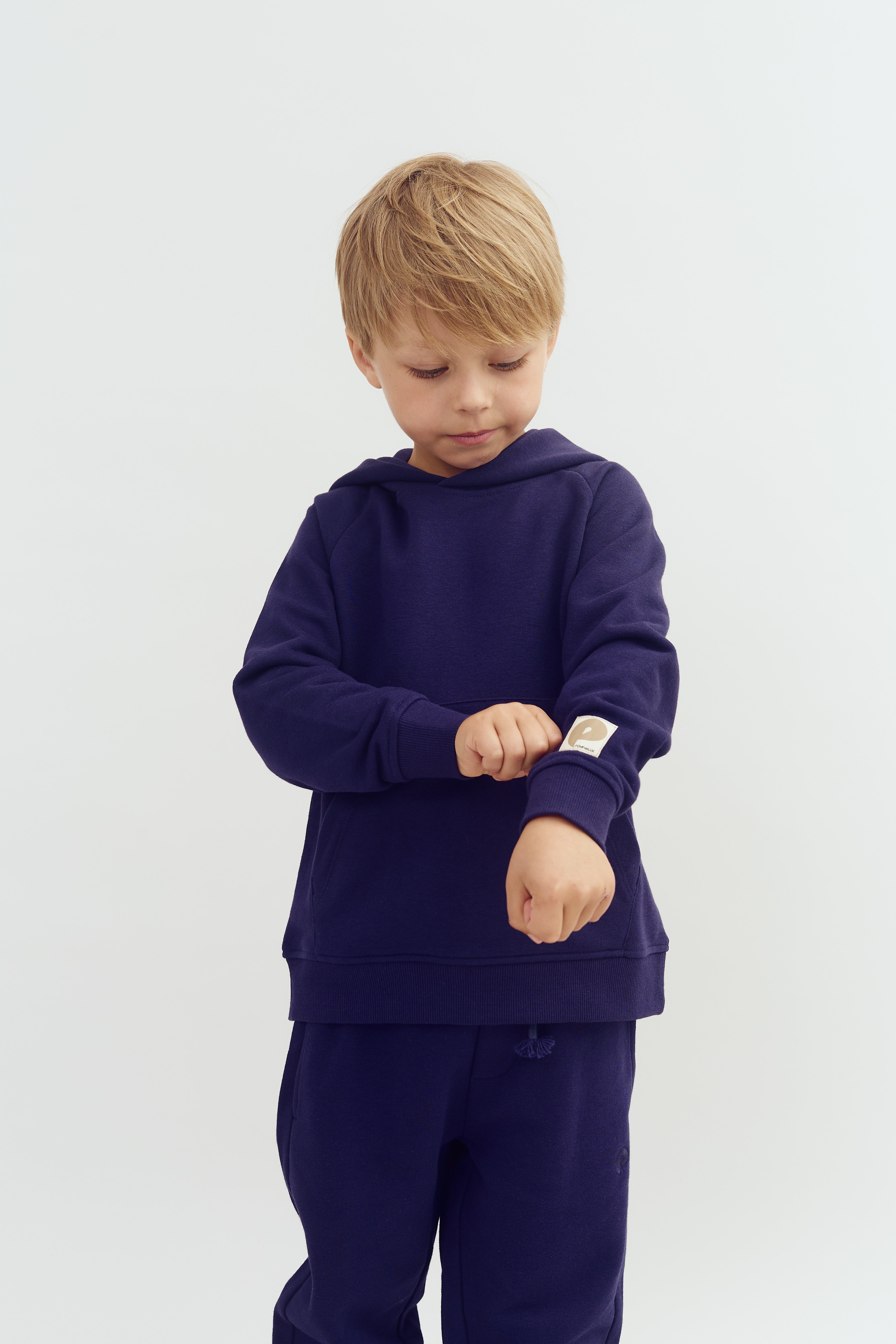 A child wearing a navy tracksuit adjusts his sleeve, standing against a plain white background. A patch with the letter "P" and the word "People" is visible on the sleeve.