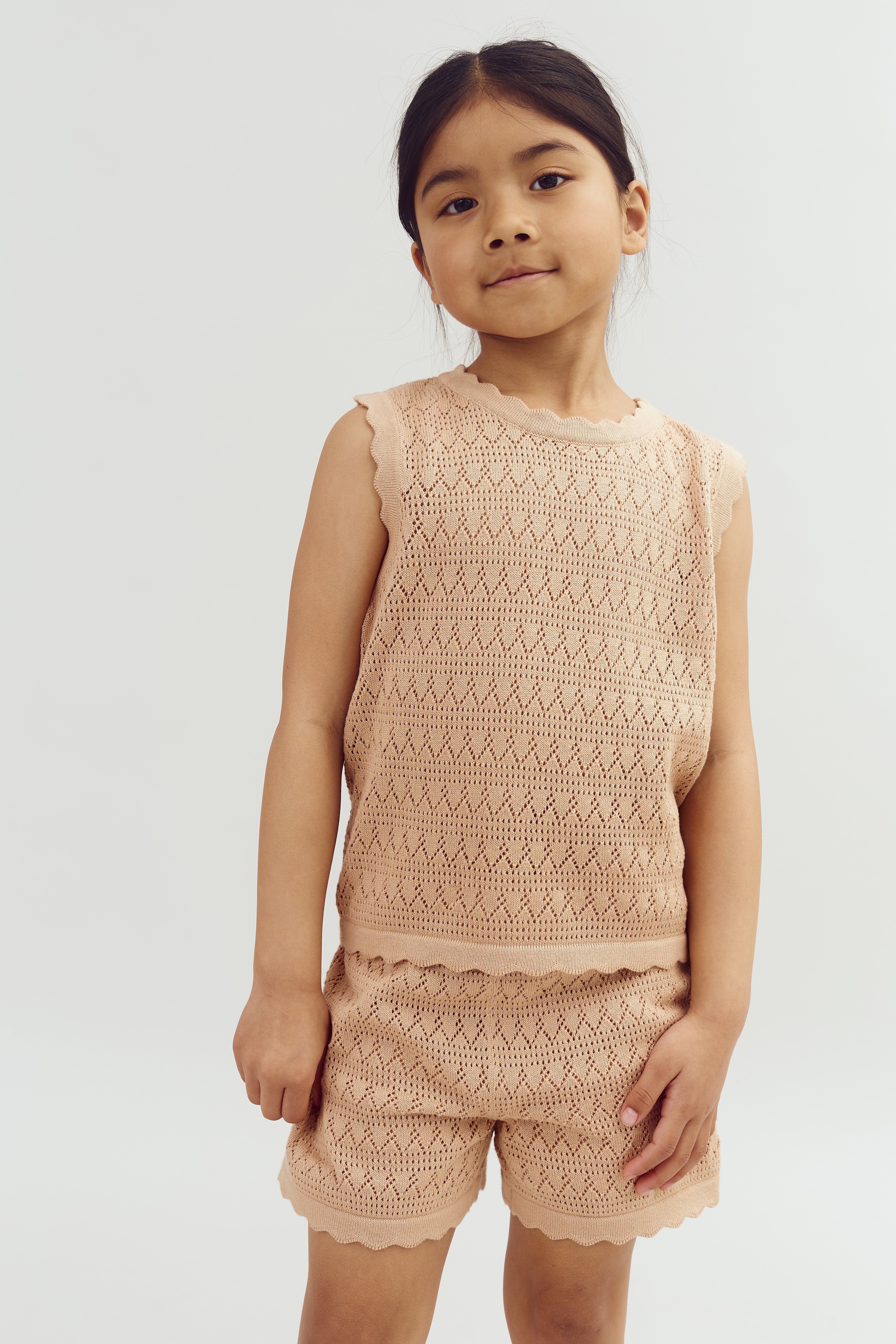 A child stands confidently wearing a light brown, sleeveless crochet top and matching shorts, against a plain white background.