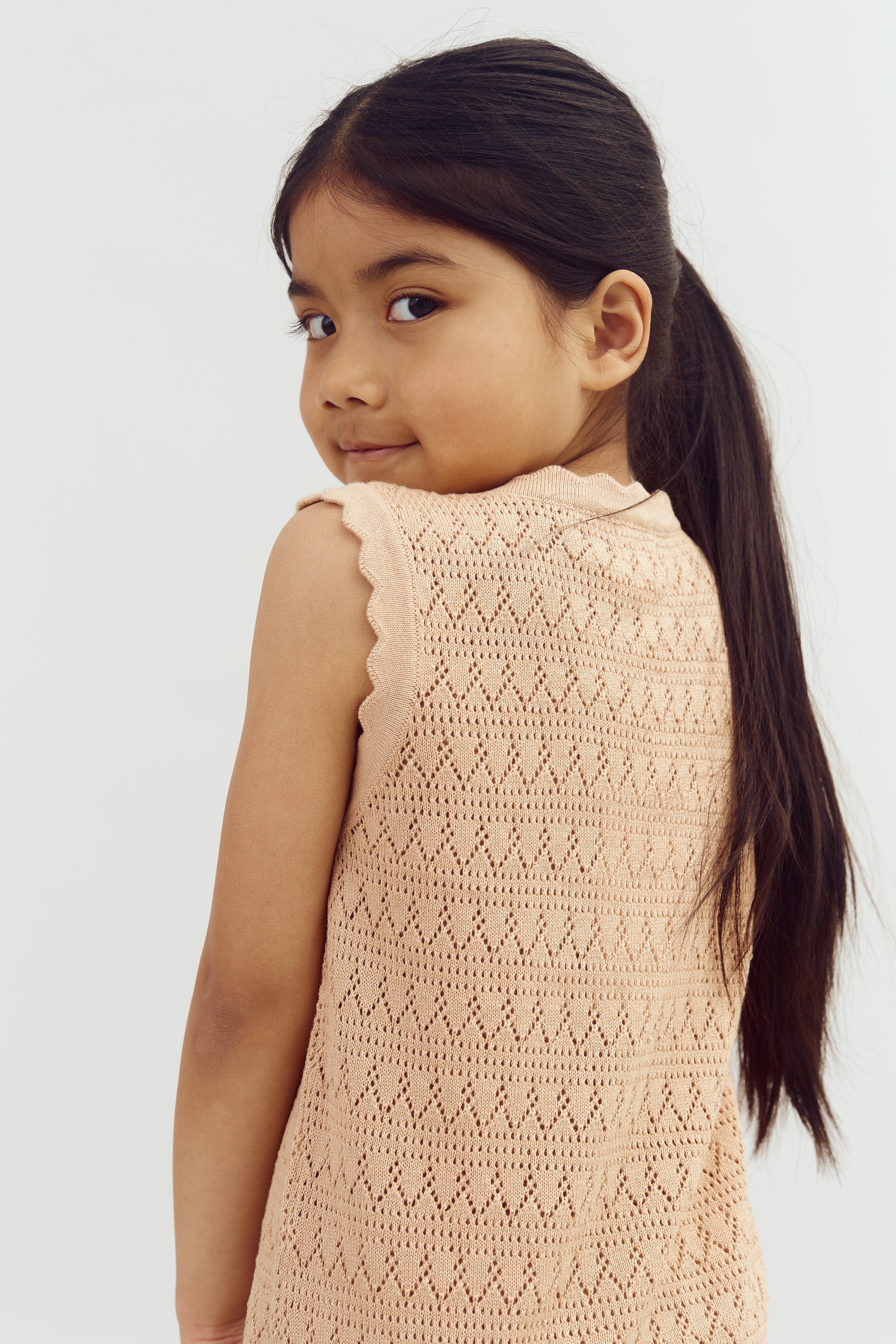 A child with long, dark hair looks over their shoulder, wearing a peach knitted top, against a plain white background.