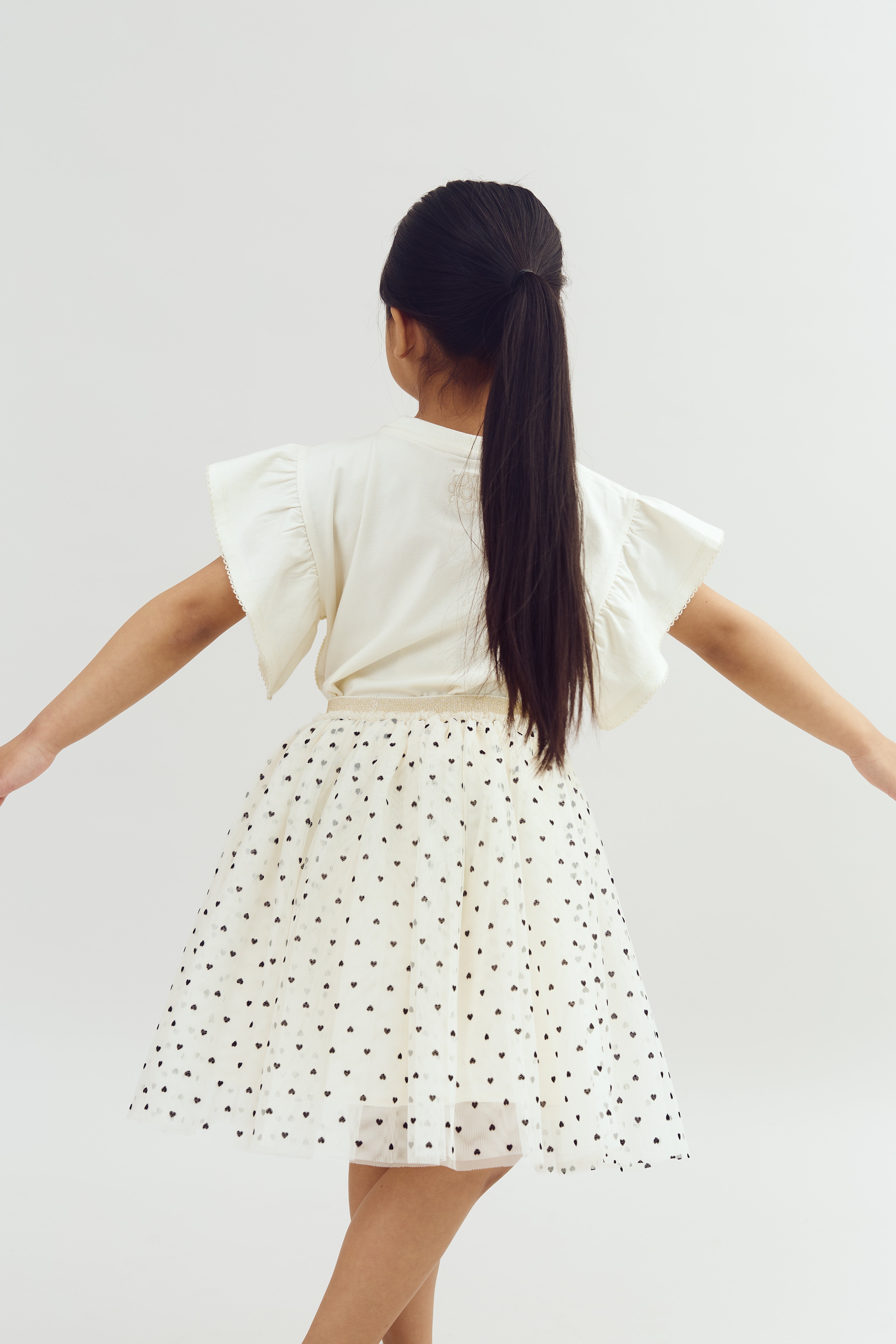 A girl stands with arms outstretched, wearing a white dress with black polka dots, in a neutral, light-colored background.
