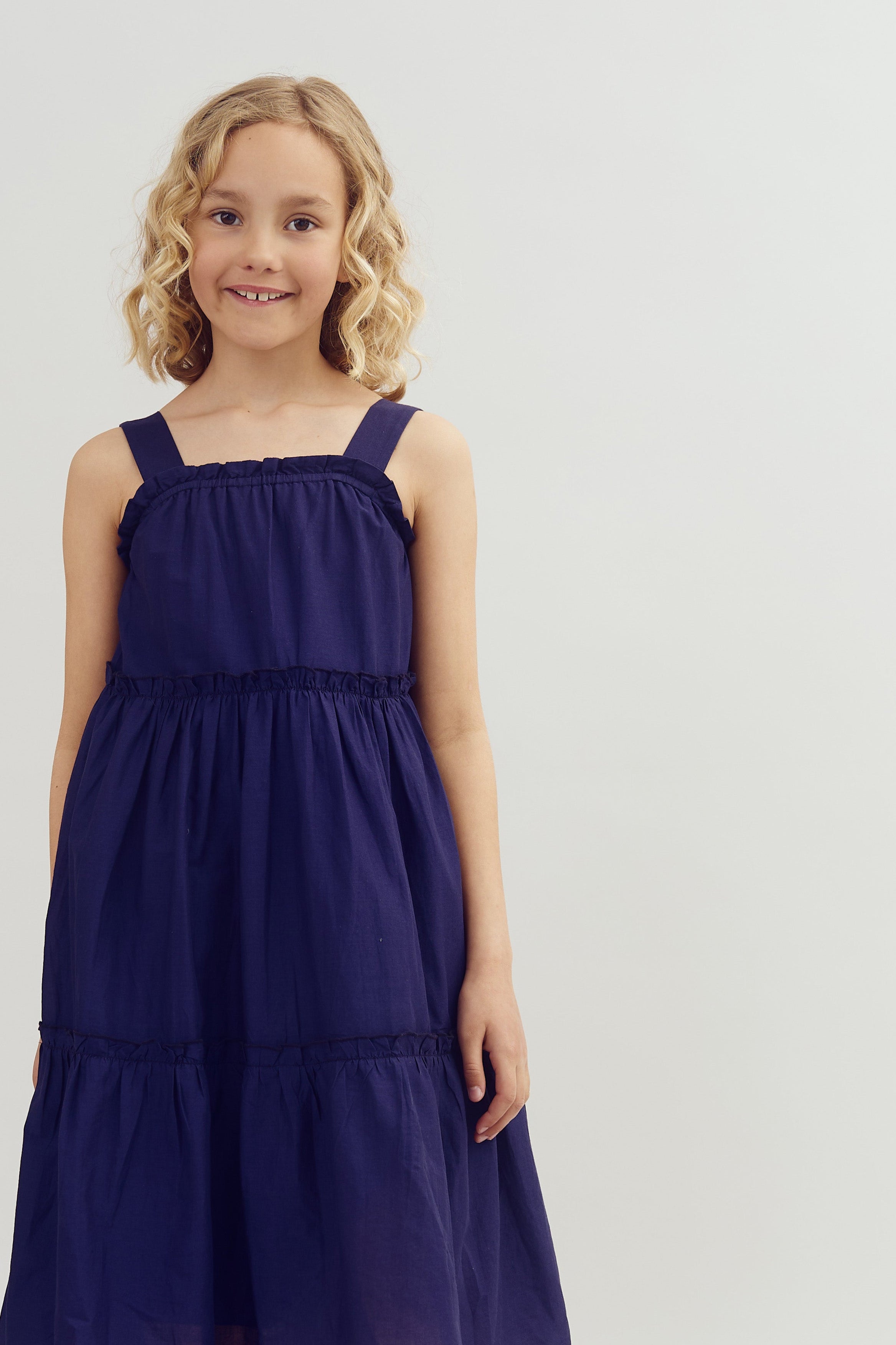 A young girl stands smiling, wearing a navy blue sleeveless dress with ruffled details, in front of a simple, light gray background.