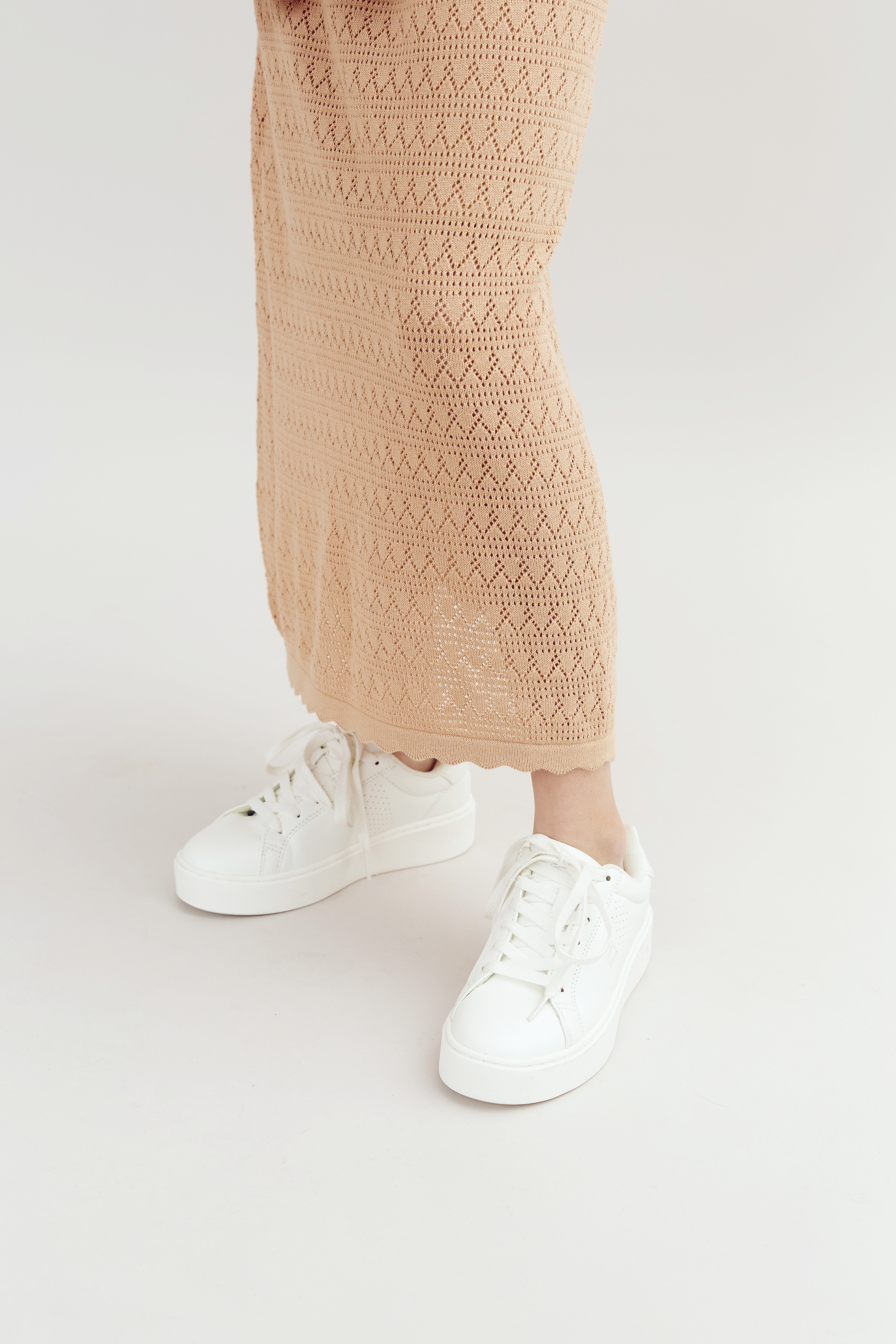 White sneakers are worn by a person standing on a light background. The shoes are paired with a tan, crocheted skirt, creating a casual and clean look.