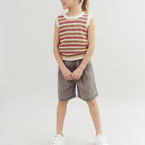 A child stands smiling, wearing a striped sleeveless top, gray shorts, and white sneakers, against a plain gray background.