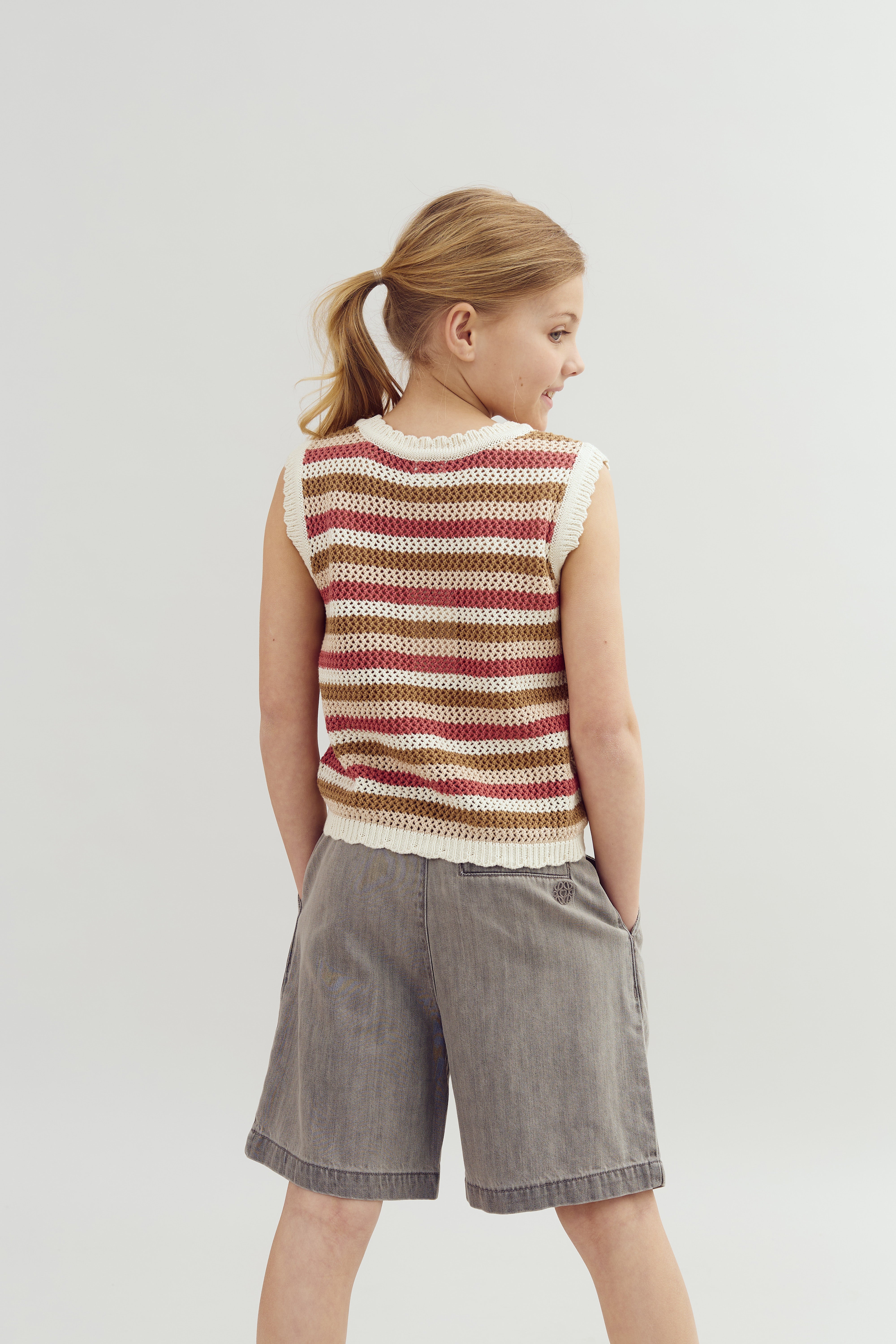 A child with a ponytail stands wearing a striped sleeveless top and gray shorts, looking to the side in a bright studio setting.
