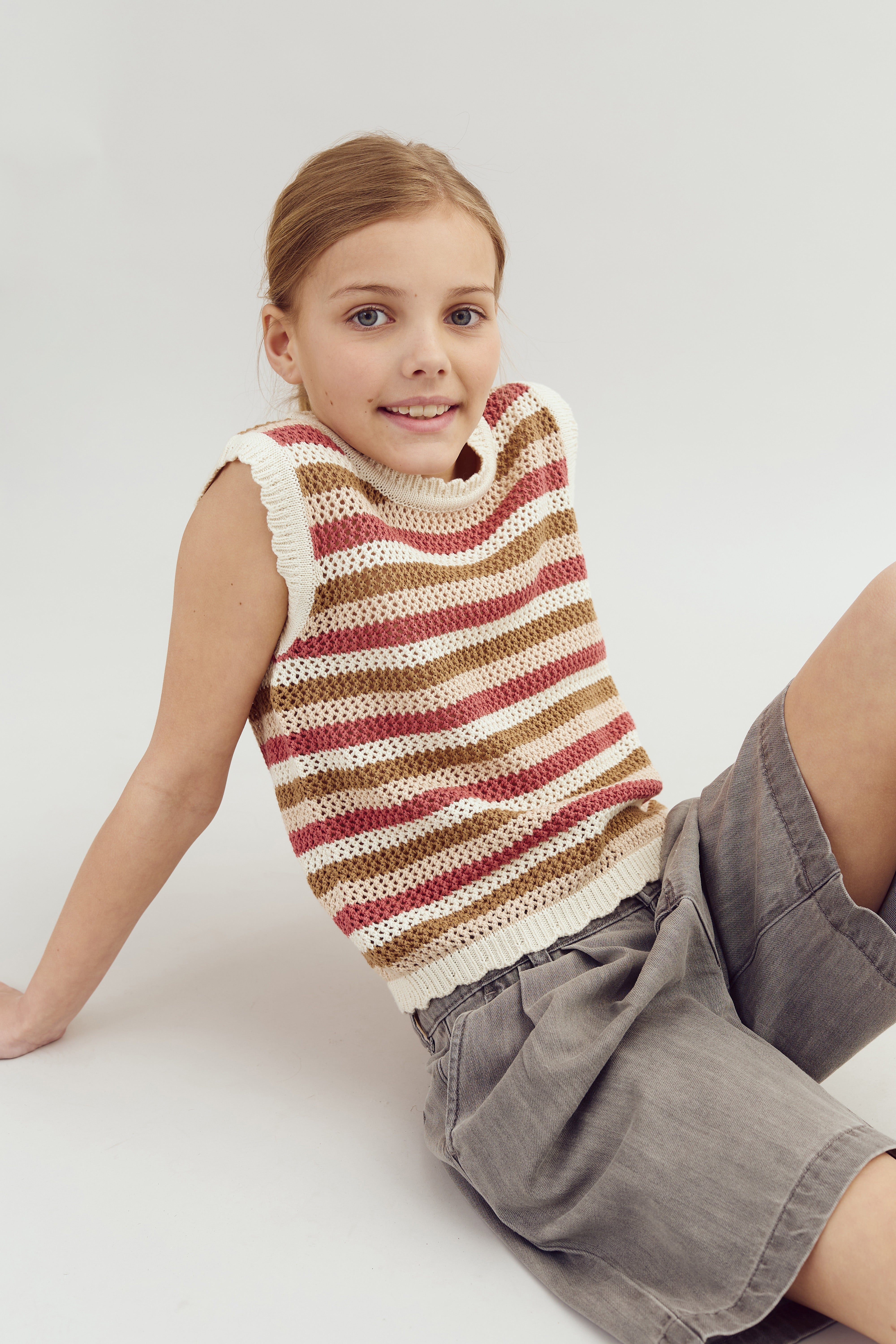 A child is seated on the floor wearing a sleeveless, striped knit sweater and gray shorts, smiling against a plain, light background.