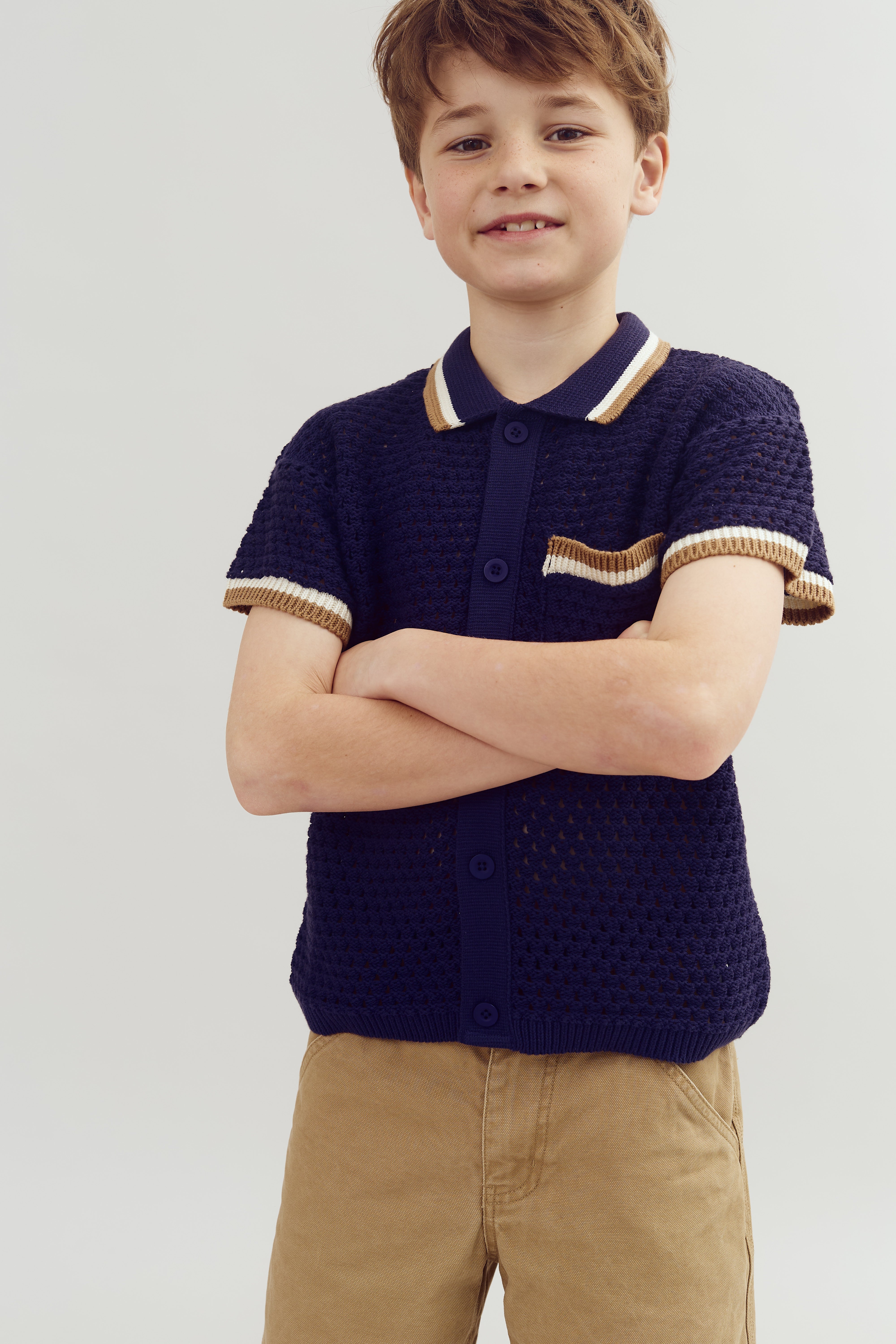 A child crosses arms while smiling, wearing a navy crocheted shirt with beige and white trim, against a plain gray background.