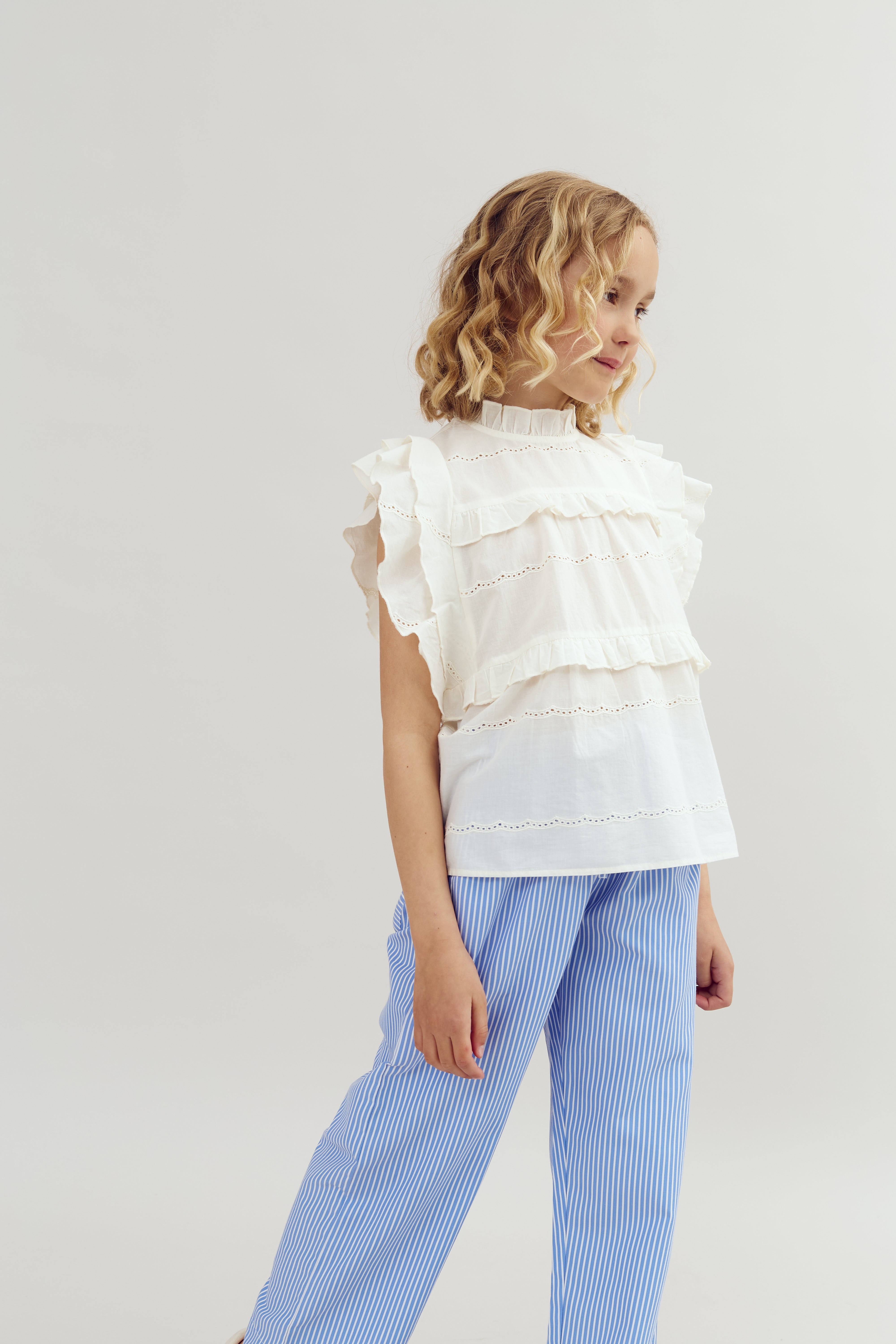 A young girl stands wearing a white ruffled blouse and blue striped pants, looking to the side against a plain, light-colored background.