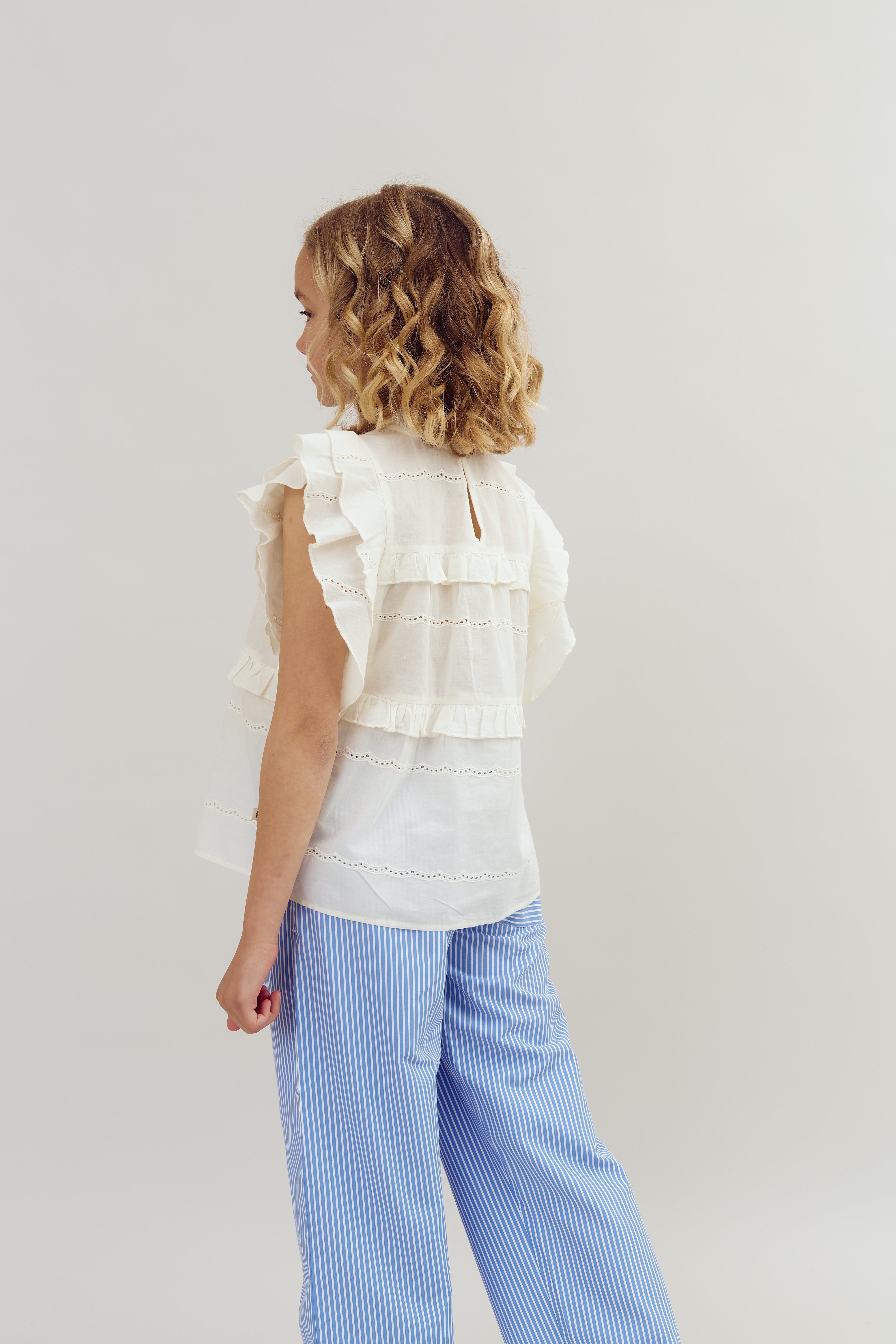 Child with curly hair is standing, wearing a white ruffled blouse and blue striped pants, against a plain light background.