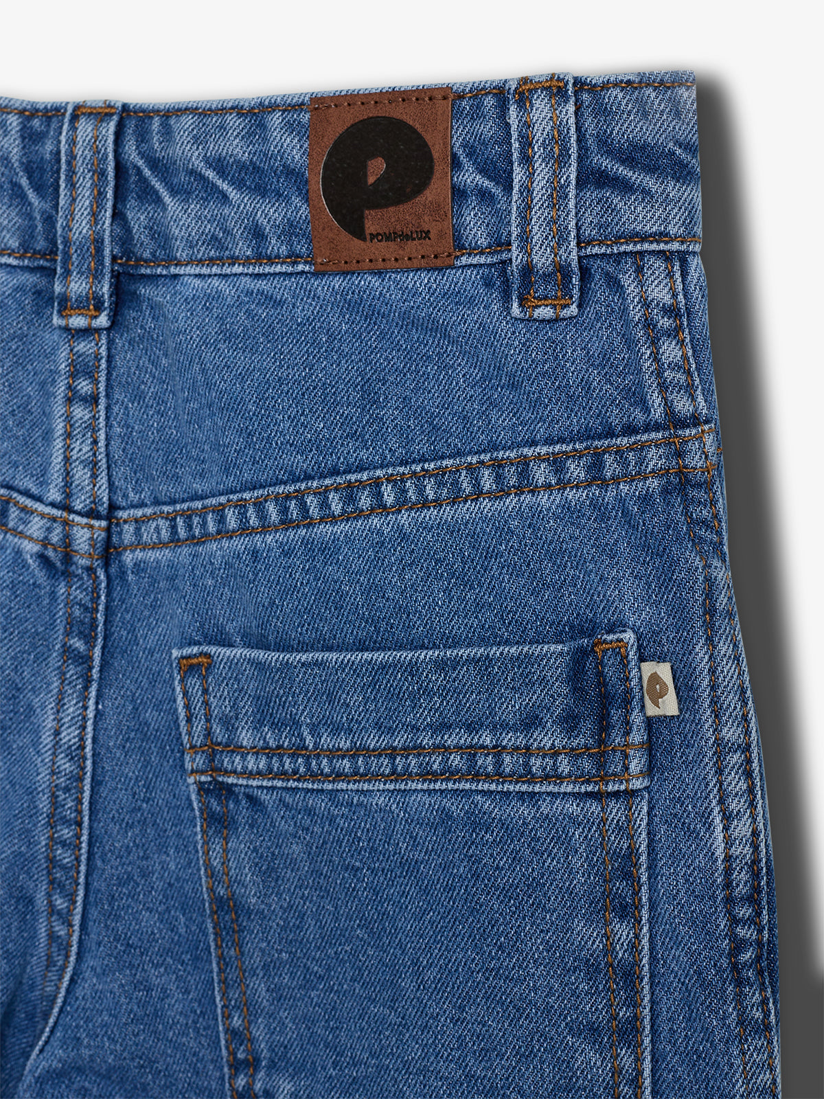 Blå denim bukser vises uden handling, set bagfra. Et brunt mærke på linningen har teksten "POMPdeLUX". En lomme med synlig syning og etikette er i venstre side.