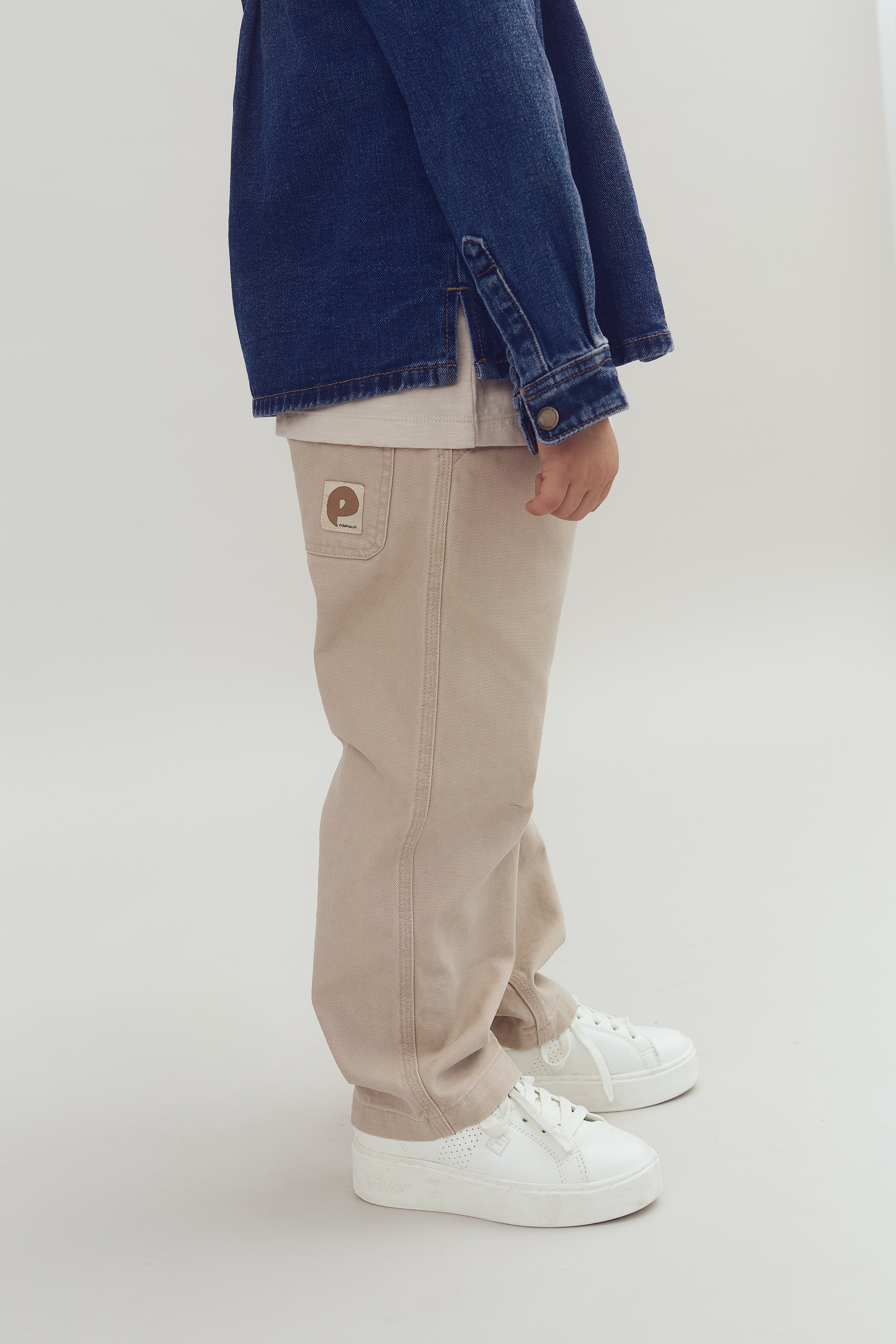 En person iført beige bukser og hvide sneakers står i et neutralt rum. En blå denimjakke dækker den øverste del. Bukserne har en mærkeplade med teksten "popeye".