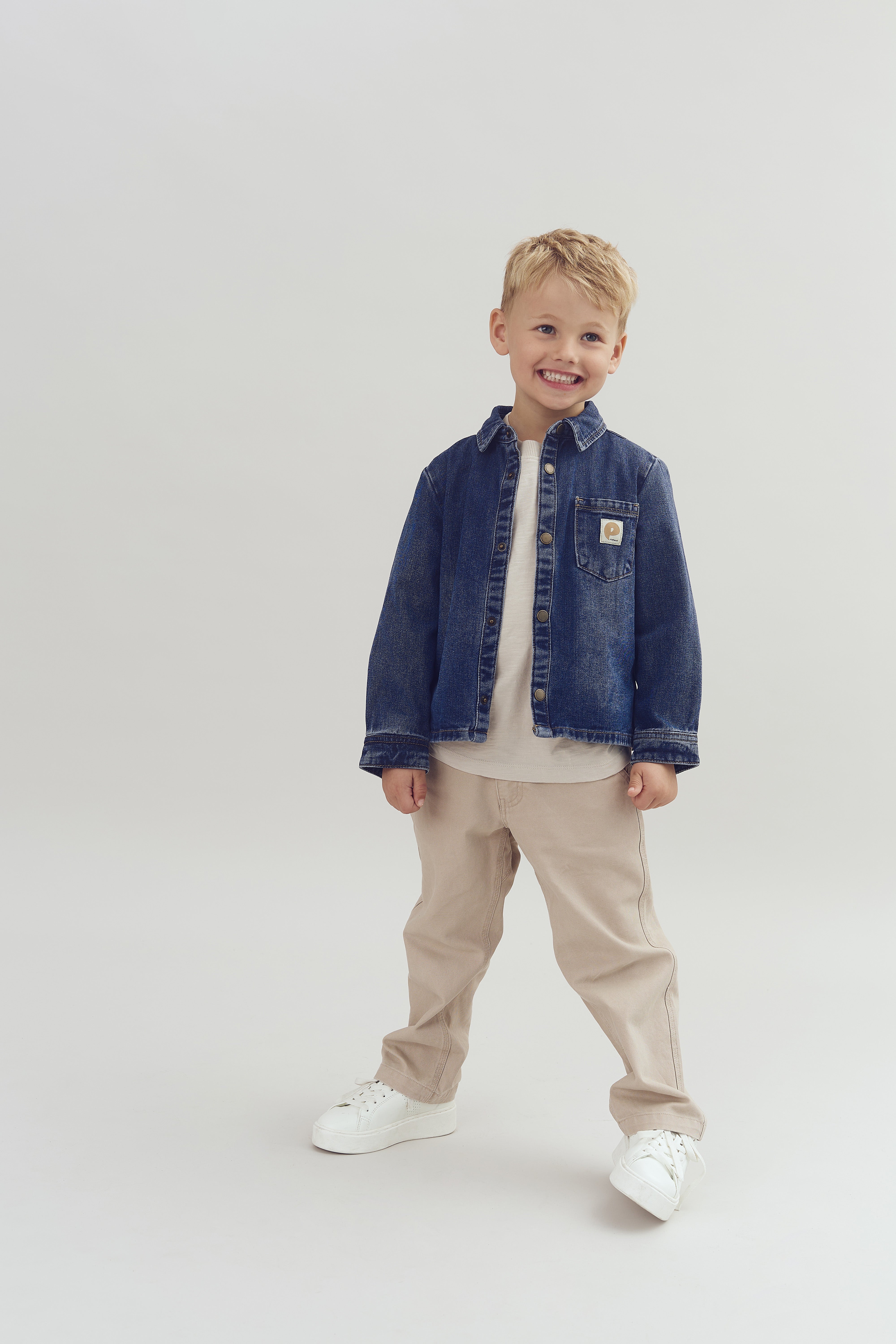 Dreng smiler og står iført en denimjakke, beige bukser, og hvide sko på en lys baggrund.