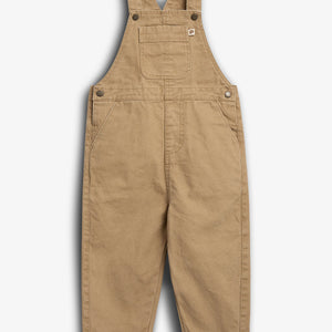Beige overall med seler hænger frit. Dets enkle design har lommer foran og synlige knapper. Baggrunden er en neutral, hvid flade, der fremhæver tøjet.