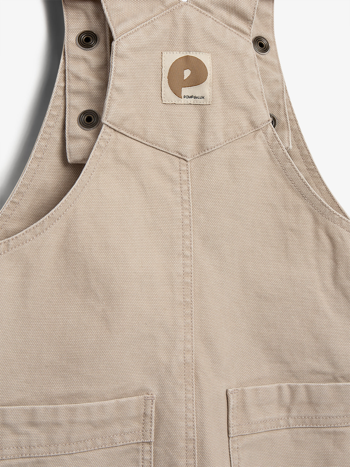 En beige overall med logoet "POMPdeLUX" påsyet foran. Der er trykknapper øverst, og overallsstykket har syede lommedetaljer. Baggrunden er hvid.