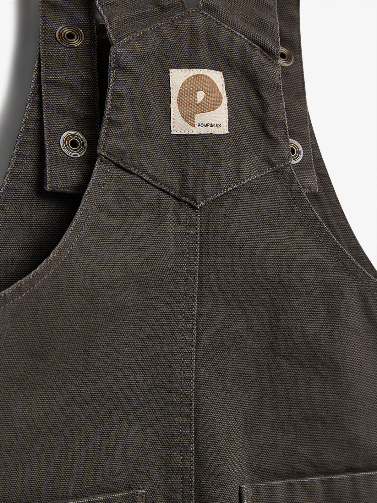 Sele med knapper sidder roligt på en grå baggrund. Et mærke med et brunt symbol og teksten "POMPdeLUX" sidder på stoffet, der har en tung tekstur.