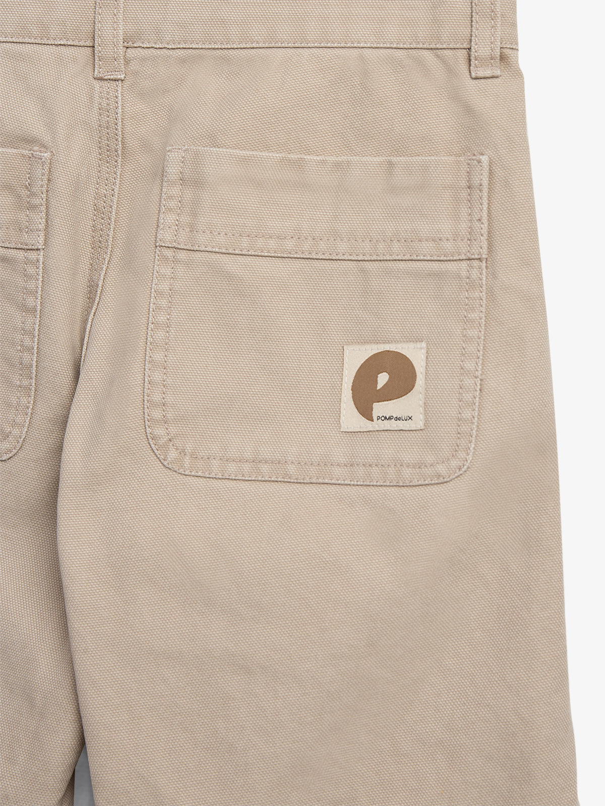 Beige bukser viser en lomme med en stofmærkat, der har et rundt brunt logo med teksten "POMPELMUX." Bukserne er lavet af kraftigt stof.