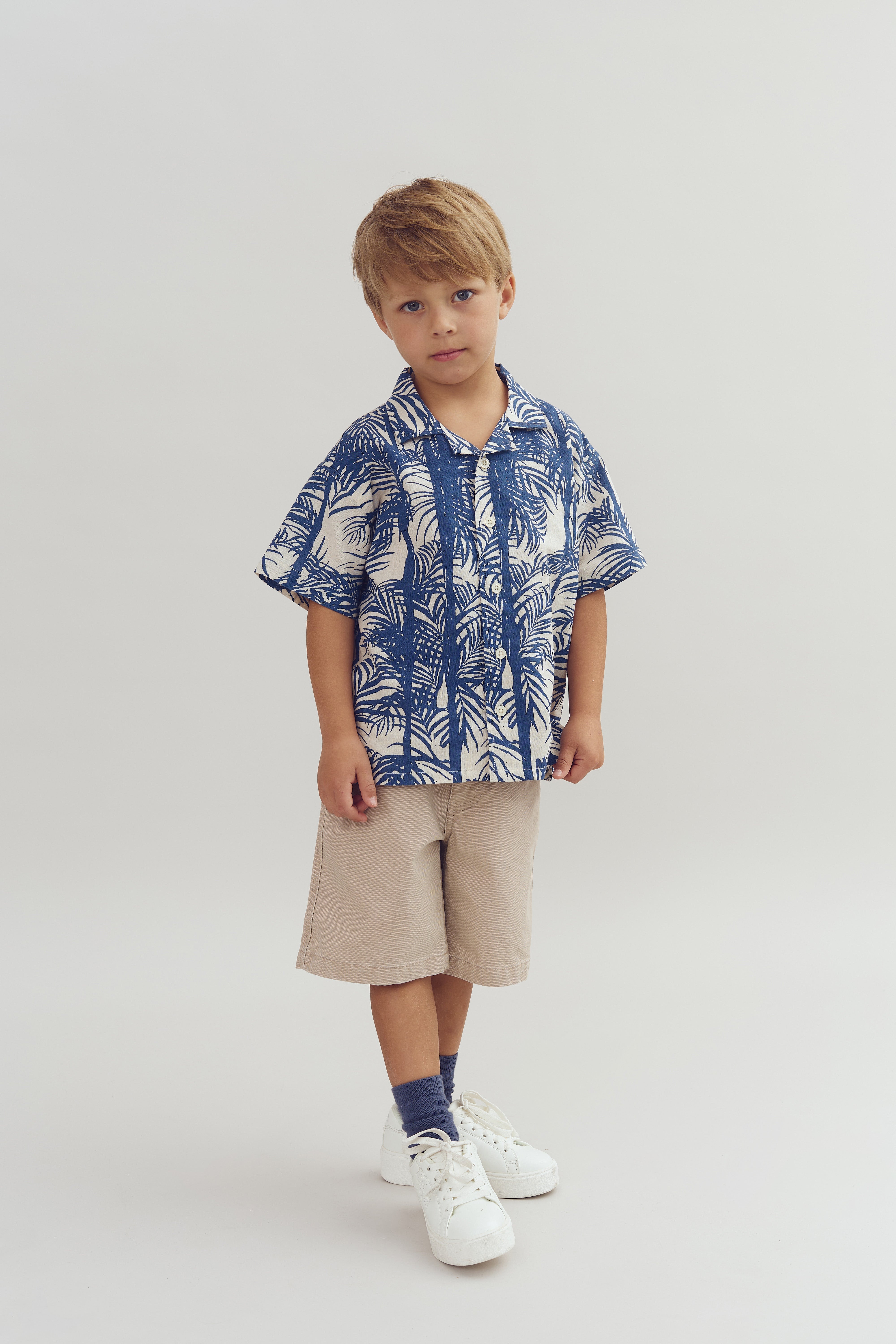 En dreng står stille iført en blå-hvid mønstret skjorte og beige shorts i en lys, neutral baggrund. Han bærer hvide sneakers og blå sokker.
