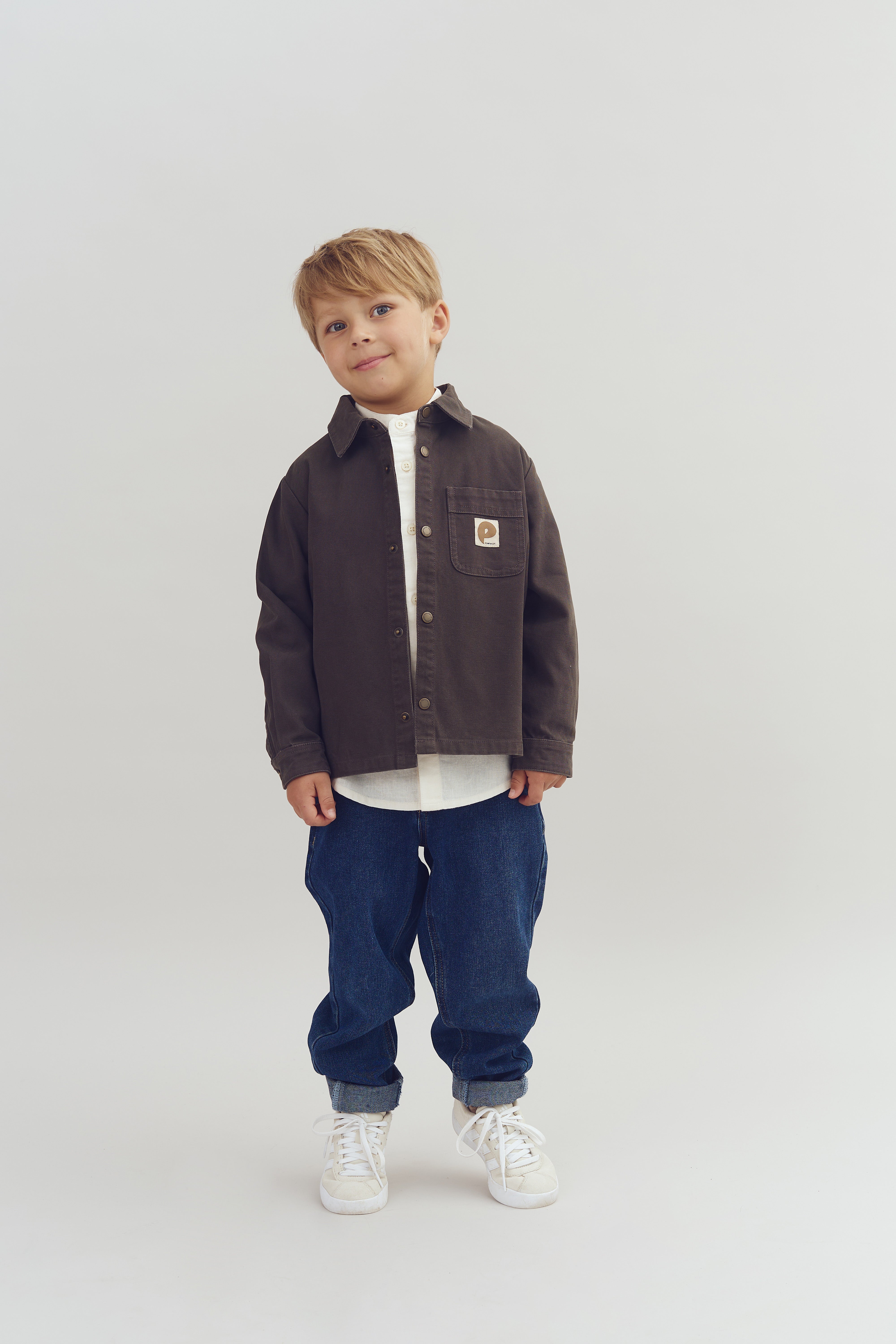 Dreng står og smiler iført en brun jakke med et logo på lommen, hvide sneakers og blå jeans; baggrunden er ensfarvet lys.